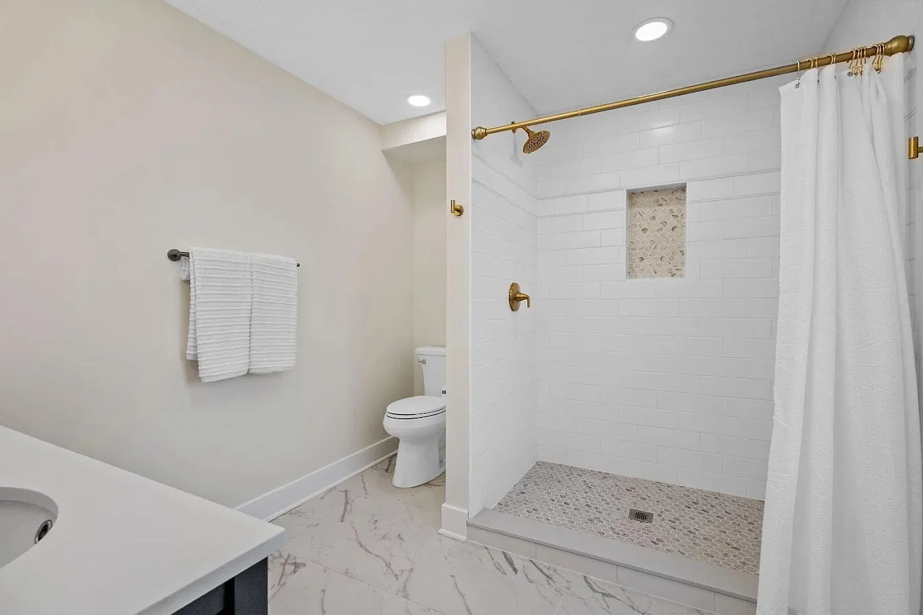 Bathroom with a white tile walk-in shower with gold fixtures and a pebble floor, white marble floor, and a toilet next to the shower.