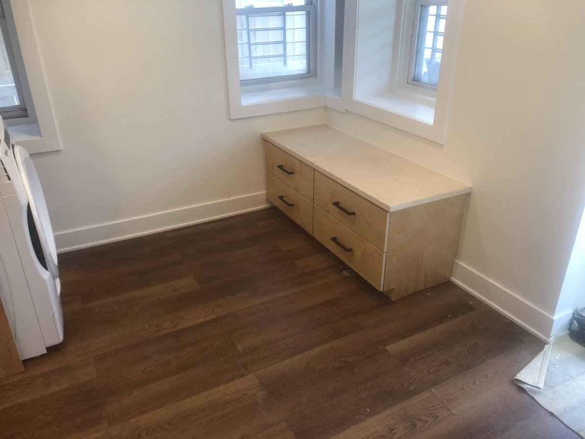 Room with wooden floor, white walls, a wooden cabinet with four drawers under two windows, and part of a washing machine on the left.