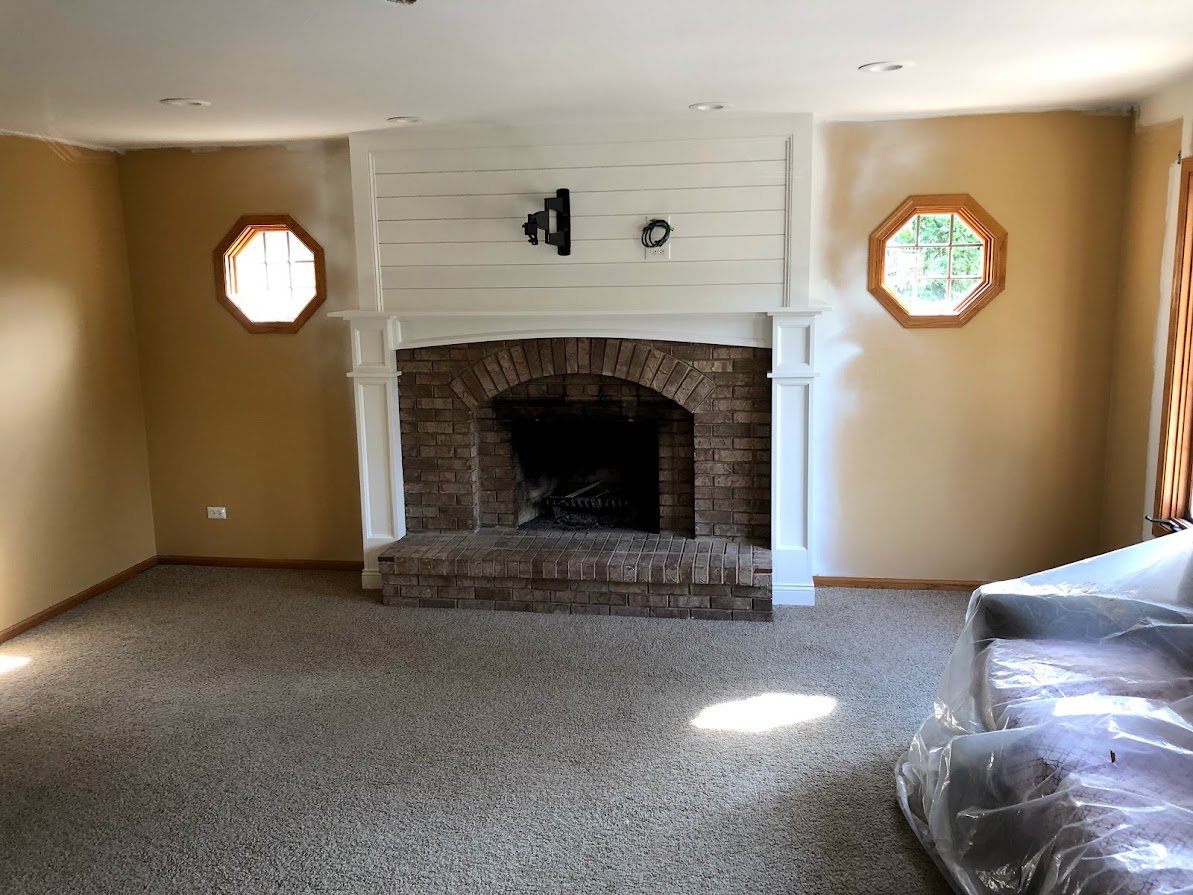 Living room with brick fireplace, wooden trim, two octagonal windows, beige walls, and carpeted floor, with furniture covered in plastic.