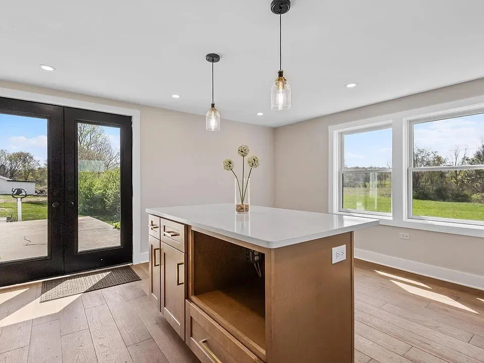 Modern kitchen with a white countertop island, two glass pendant lights, large windows, and a black glass door leading outside, with a view of a green lawn and trees.