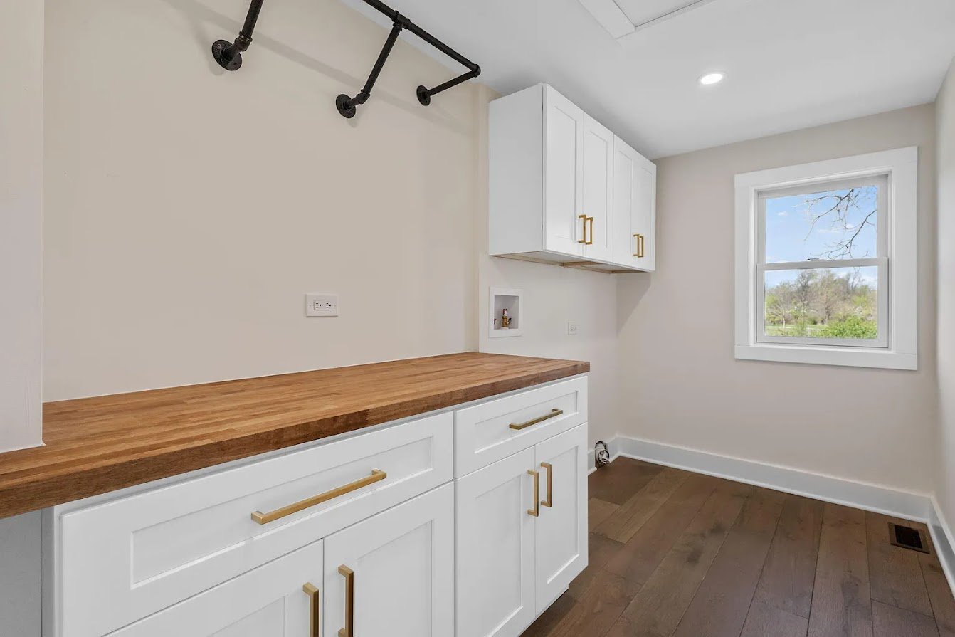 Empty laundry room with white cabinets, wooden countertop, window showing outdoor trees, and ceiling pipe fixture.