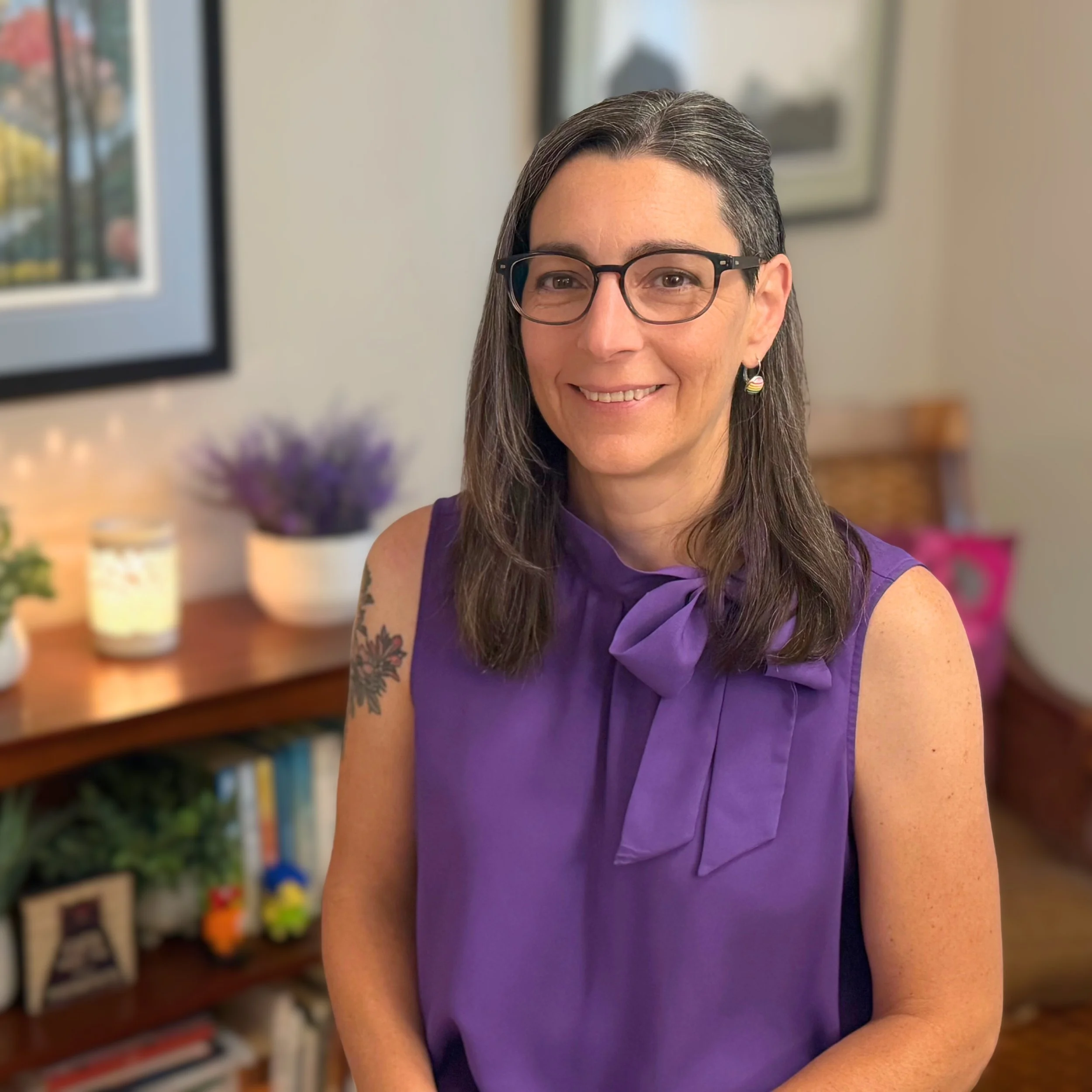 A woman with glasses, long dark hair, and a purple sleeveless blouse with a bow, standing in a living room with a bookshelf, framed pictures, and decorative items in the background.
