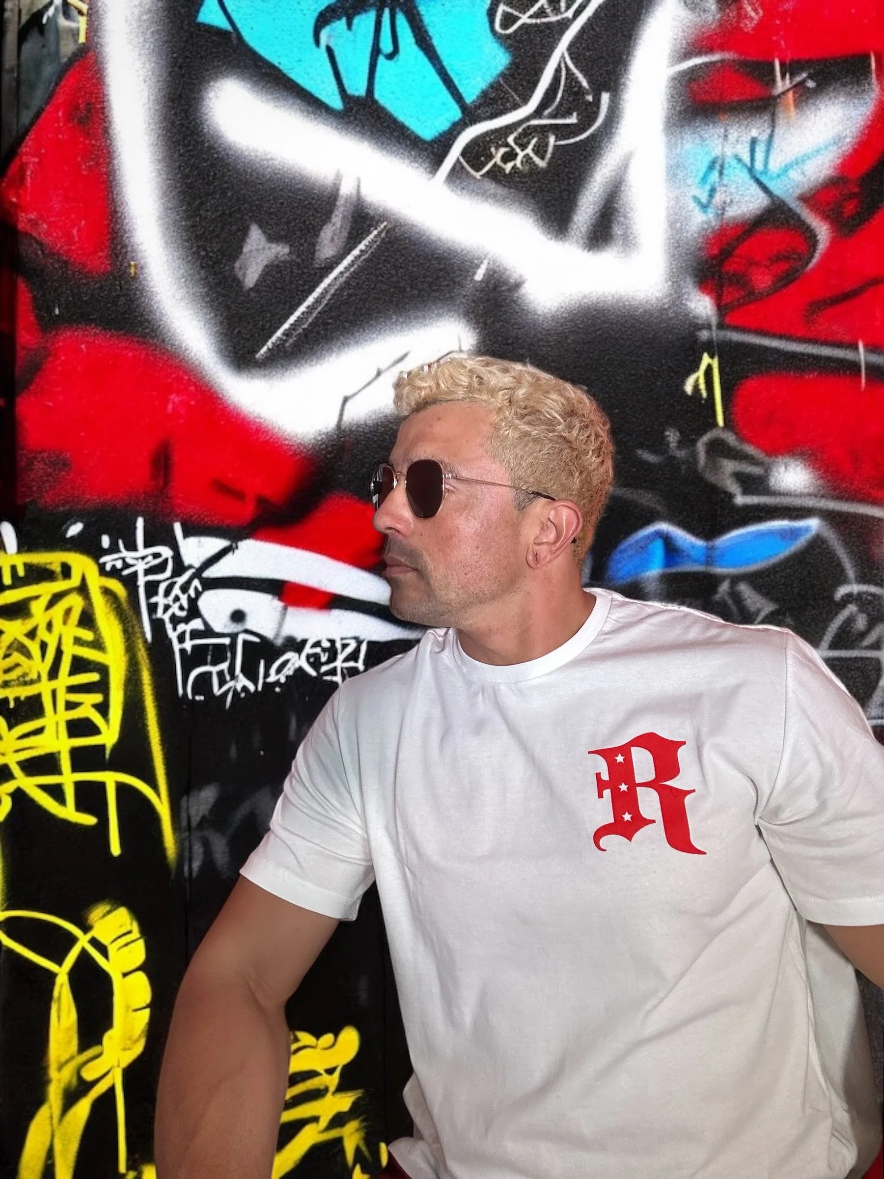 Person with sunglasses and a white t-shirt featuring a red 'R' in front of a colorful graffiti wall with abstract designs and bright colors.