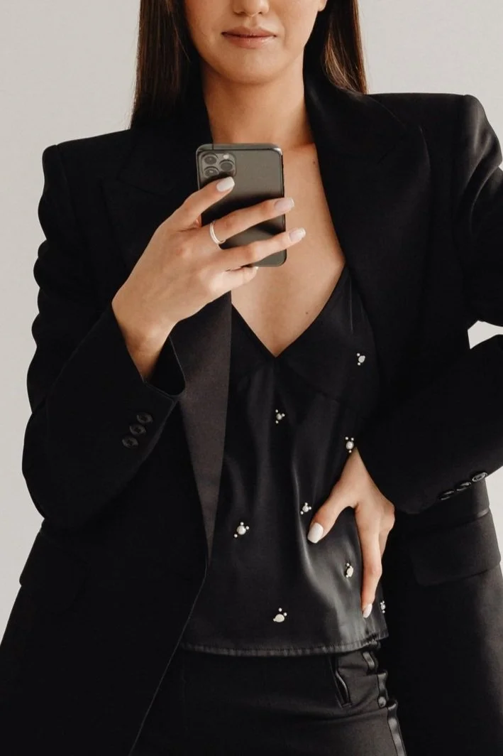 A woman taking a mirror selfie with her phone, wearing a black blazer and a black top with small pearl embellishments.