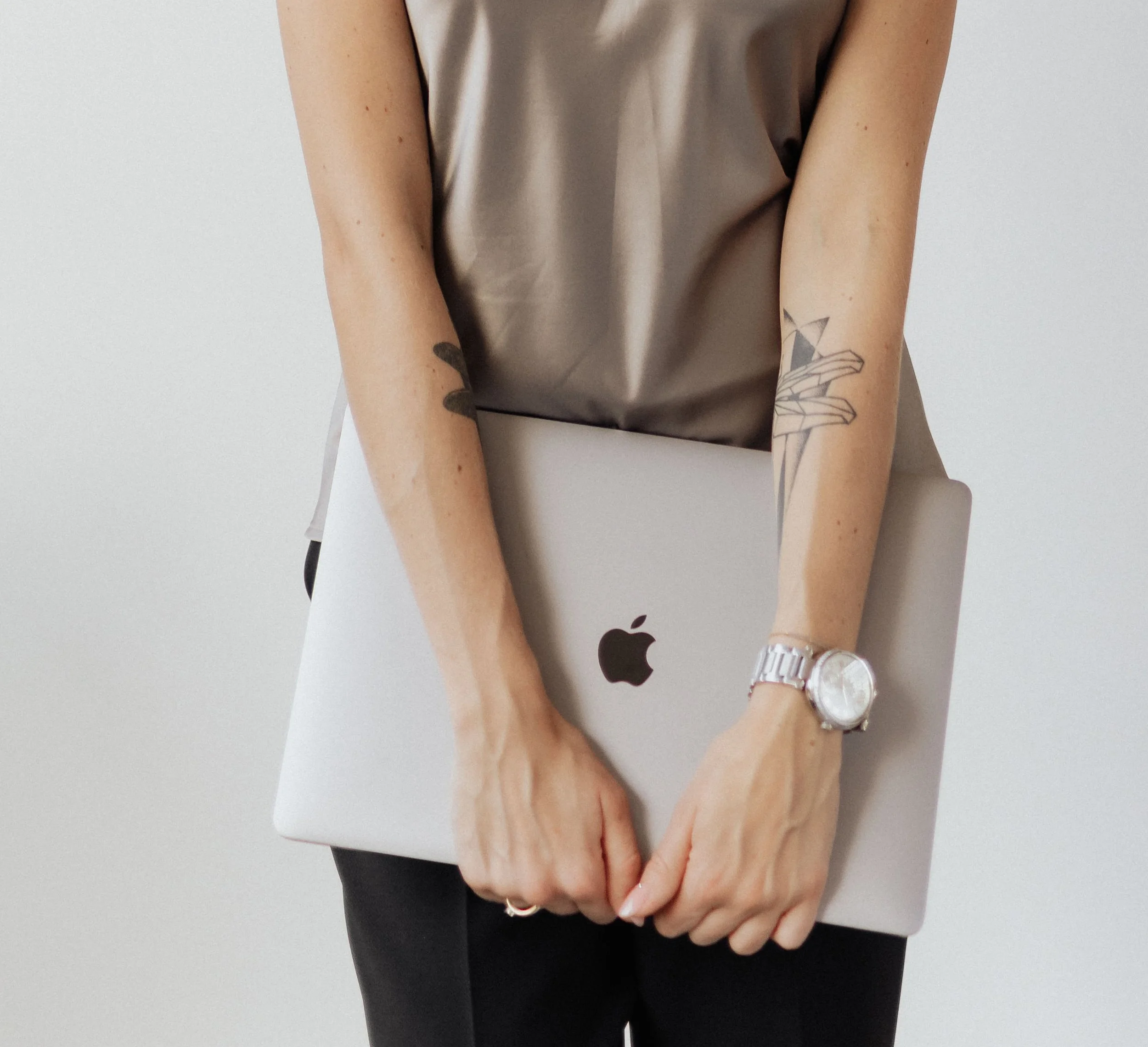 Close-up of a person holding a laptop with their left hand, showing tattoos on their forearm, wearing a beige top and silver wristwatch.
