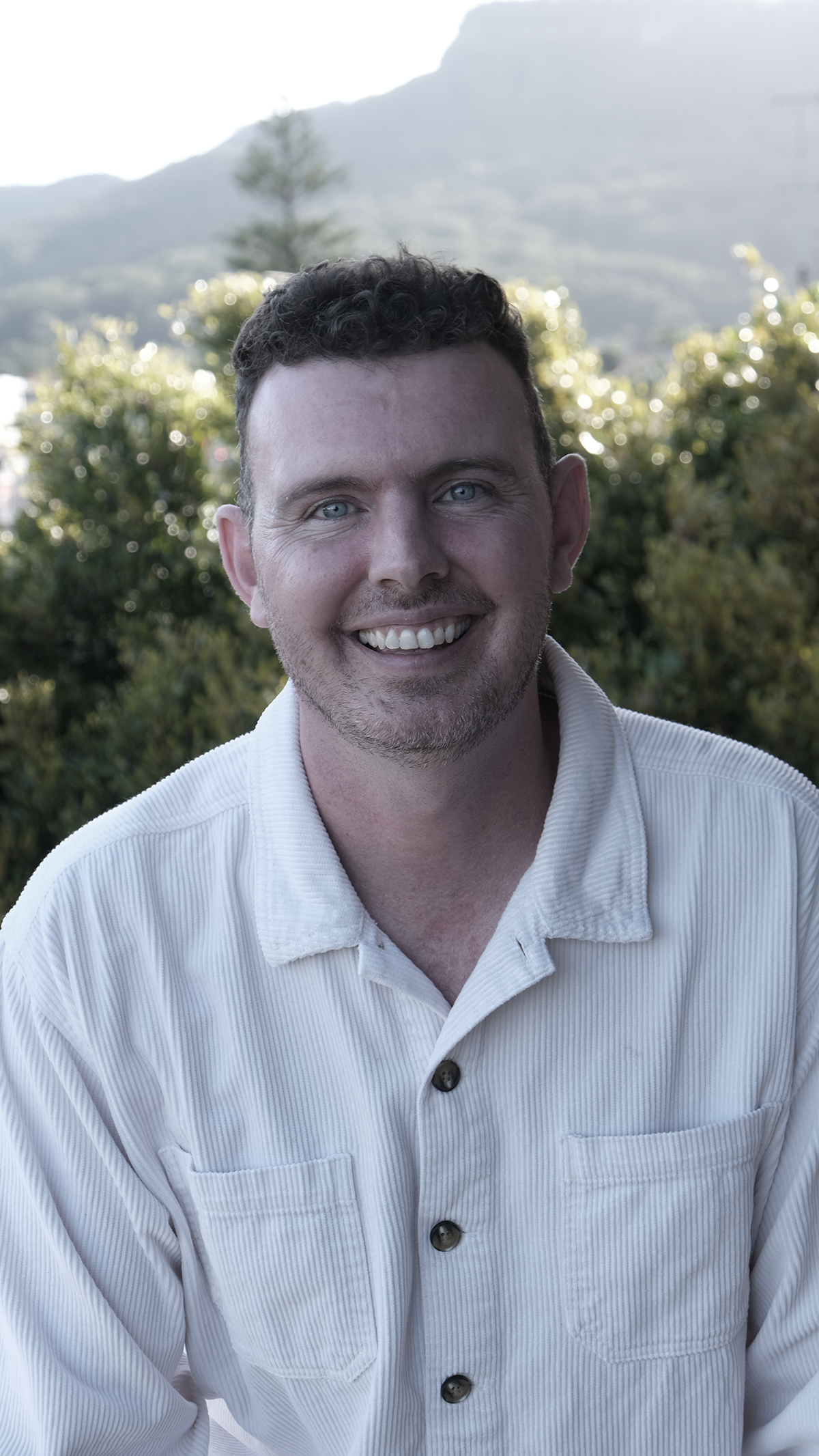Smiling person with short hair and a beard, wearing a light-colored shirt, standing outdoors with greenery and mountains in the background.