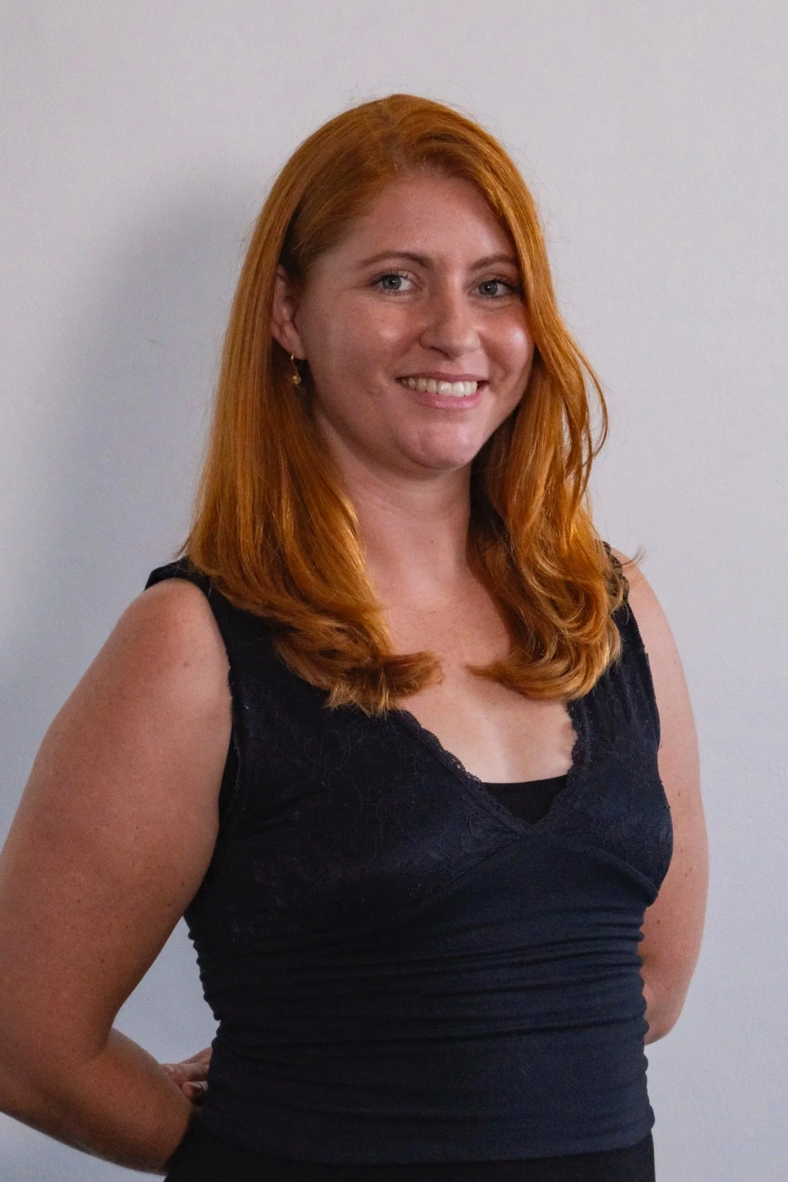 Portrait of a woman with long red hair, wearing a black sleeveless top, standing against a plain light-colored wall.