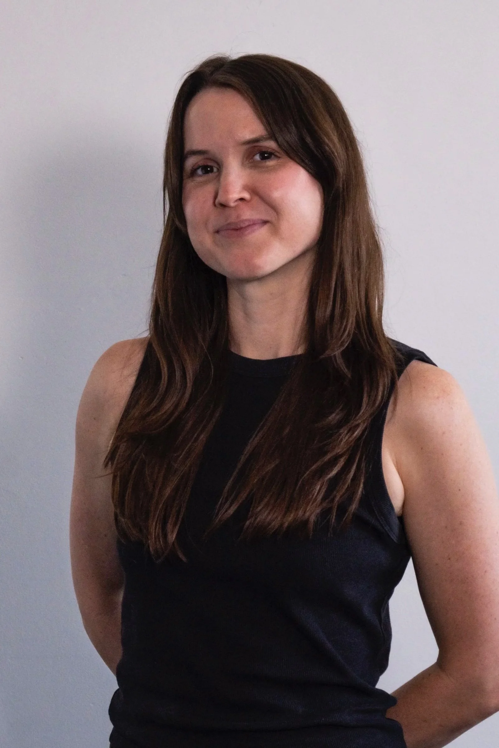 A woman with long brown hair, wearing a black sleeveless top, standing against a plain white backdrop.