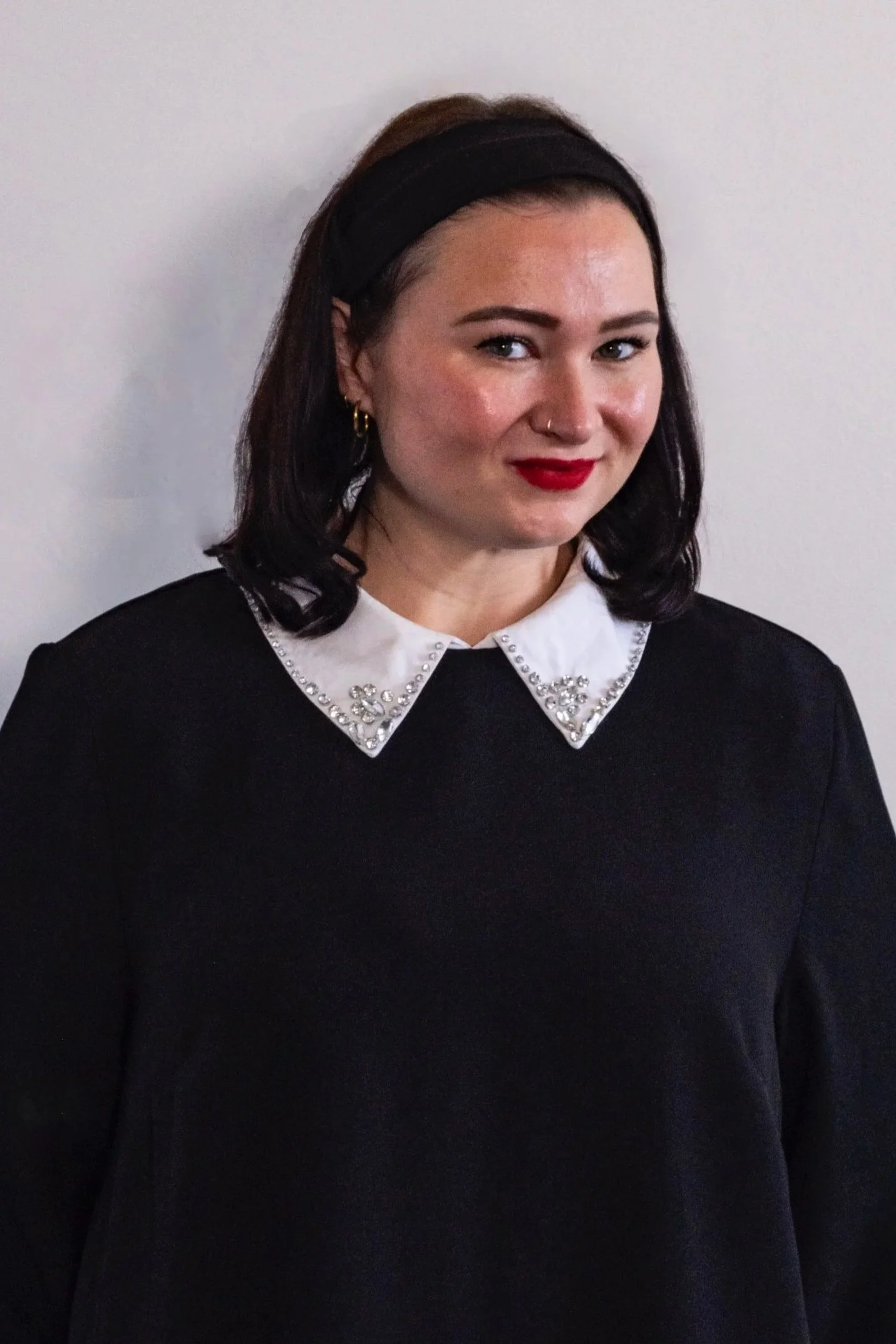 A woman with dark hair wearing a black headband, a black top with a white collar decorated with rhinestones, and red lipstick, standing against a plain white background.
