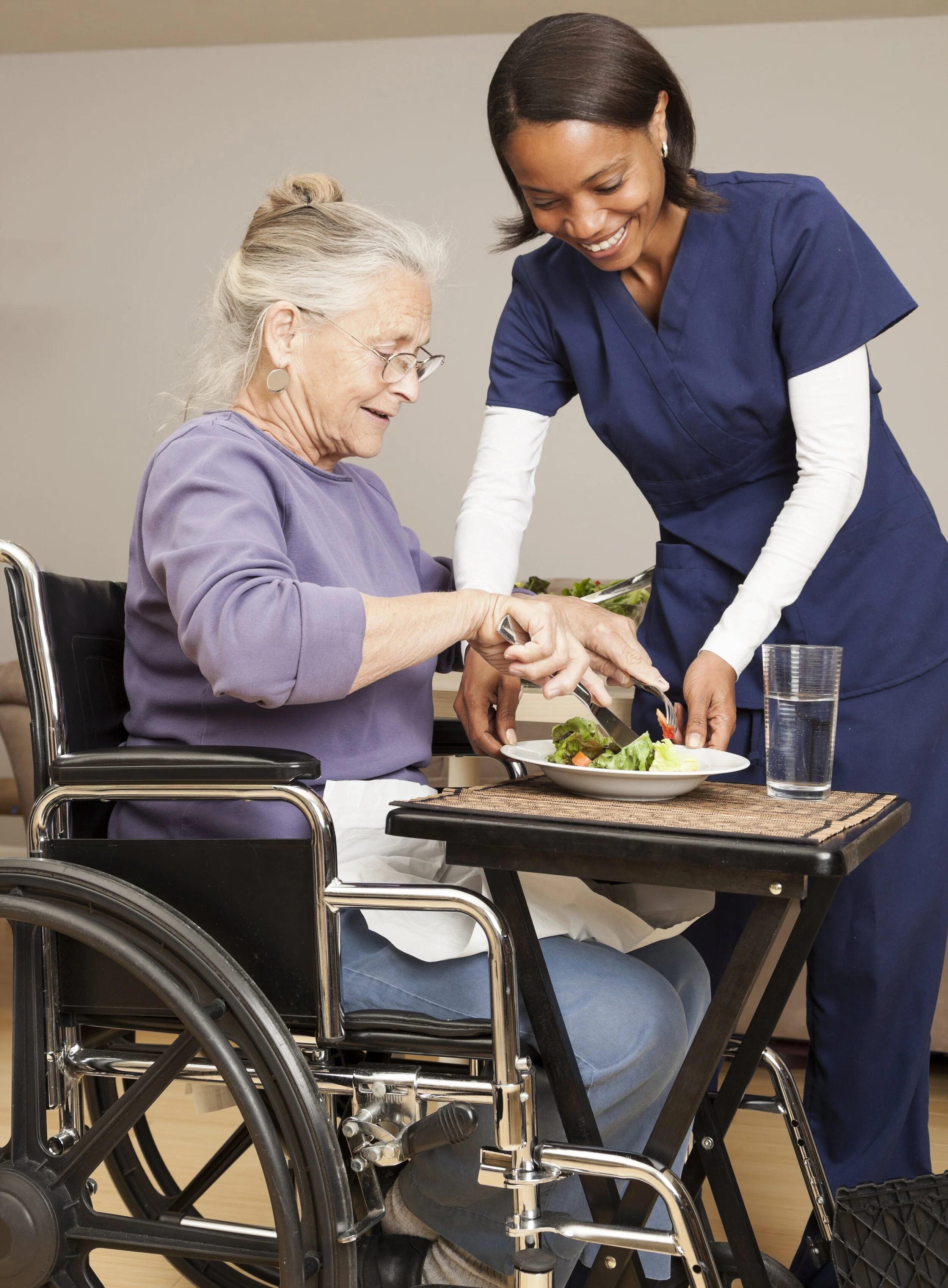 A young female caregiver helps an elderly woman in a wheelchair serve herself salad at a dining table.
