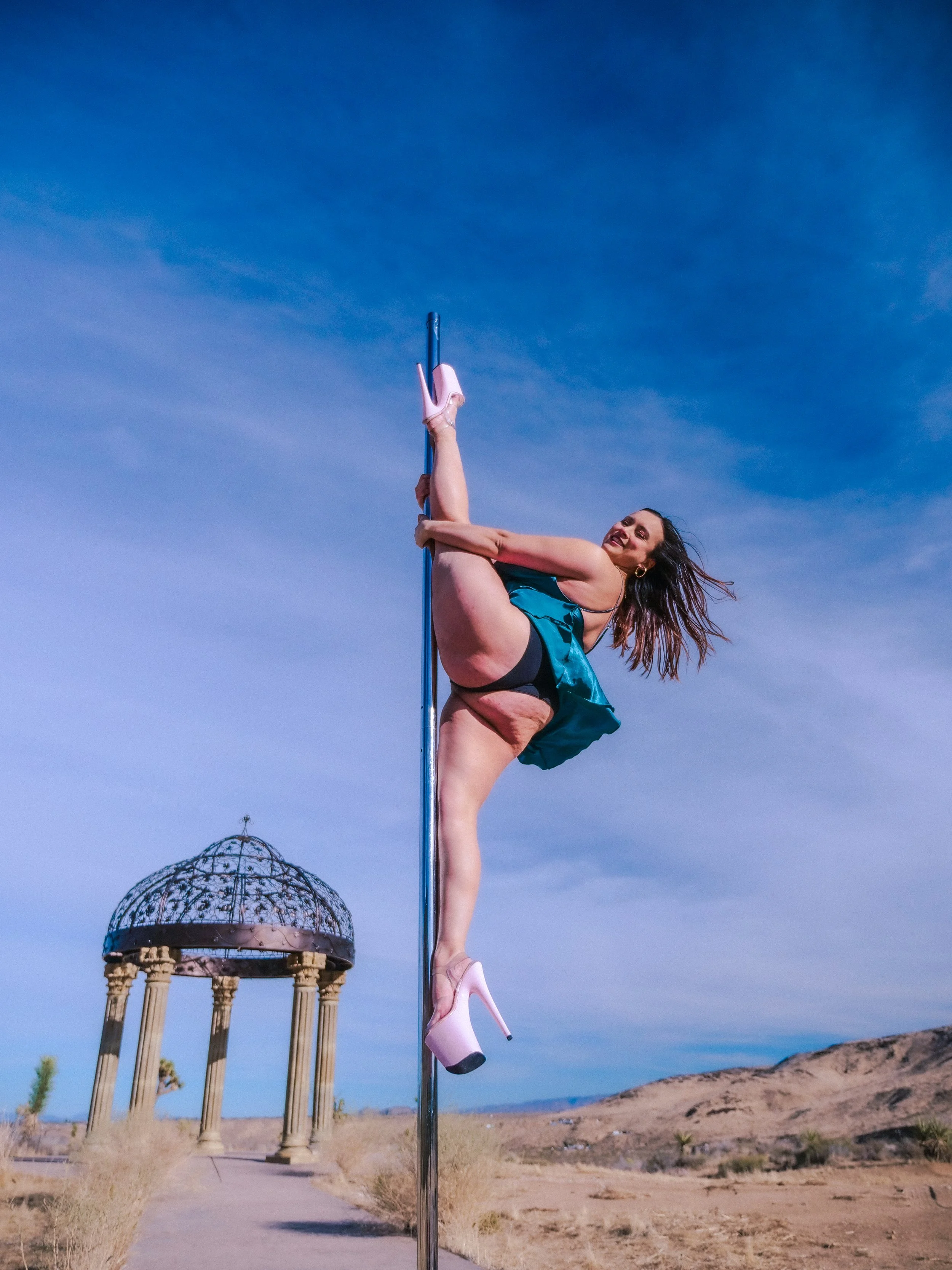 A woman in high-heeled shoes performing a pole dance in a desert landscape with a historic looking pavilion in the background.