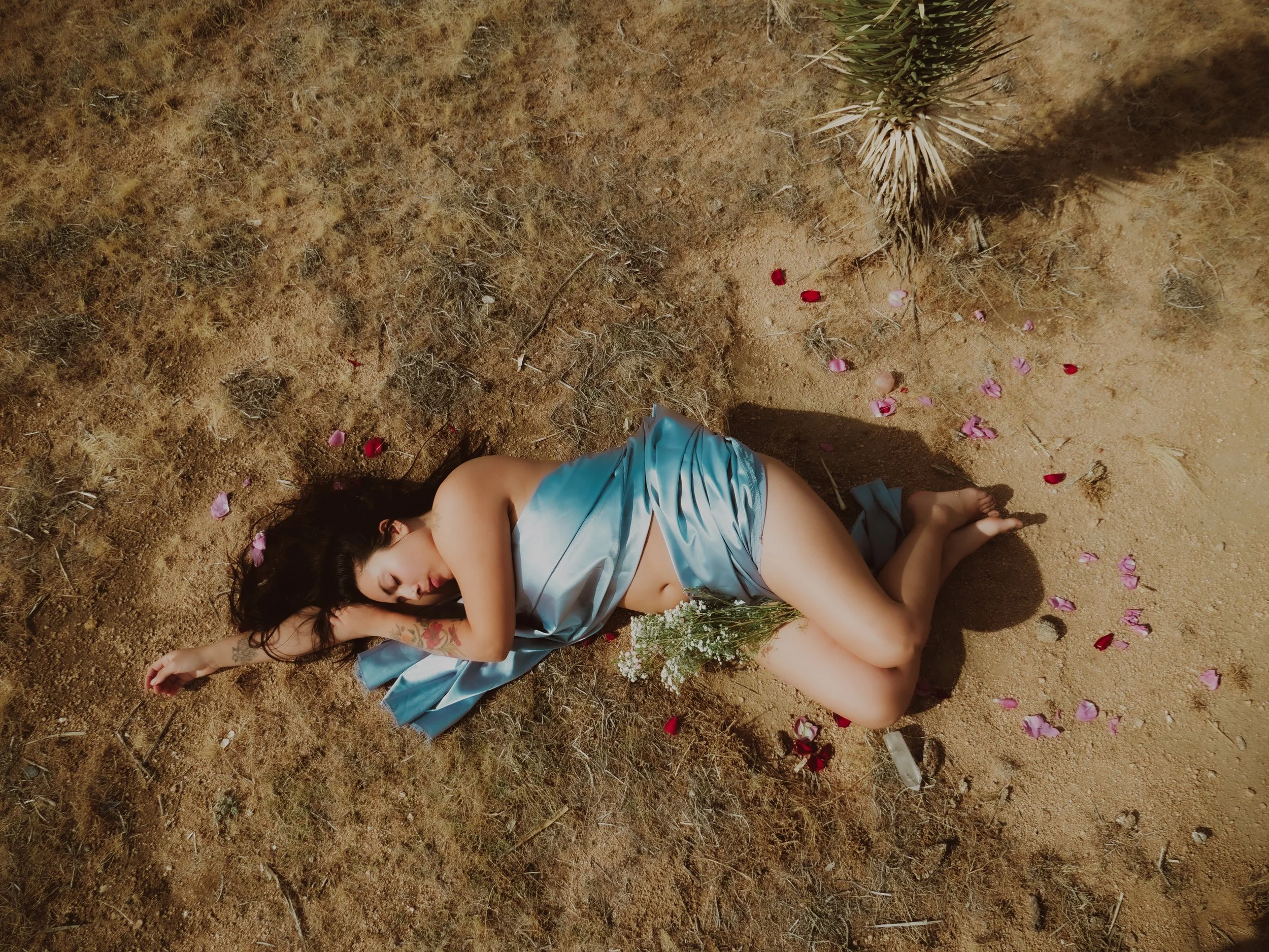 A woman wearing a blue satin dress lying on dry, sandy ground surrounded by scattered flower petals and a small palm plant.