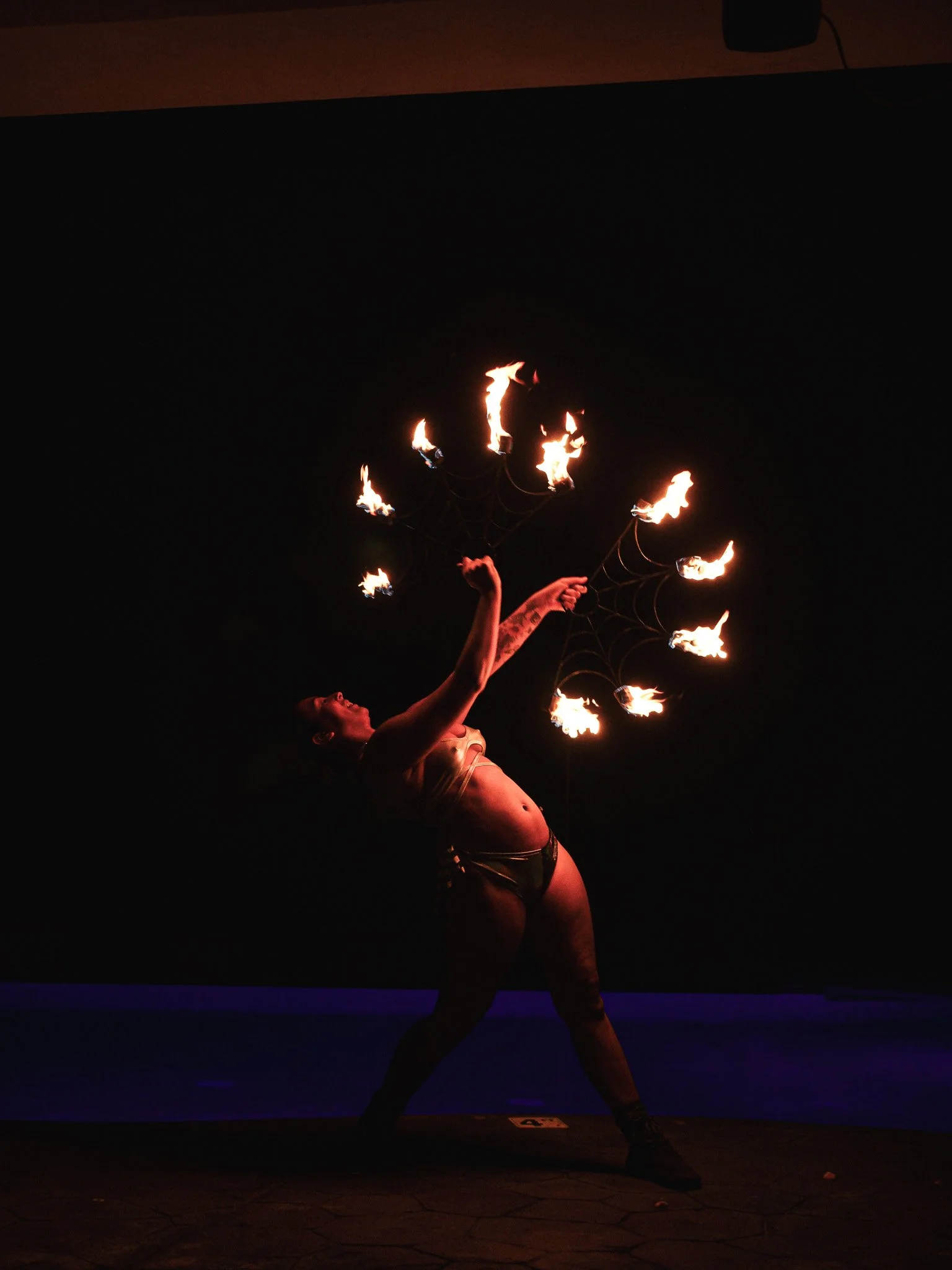 A performer in minimal clothing holding multiple flaming props with webs, creating a fiery display against a dark background during a nighttime fire dance performance.