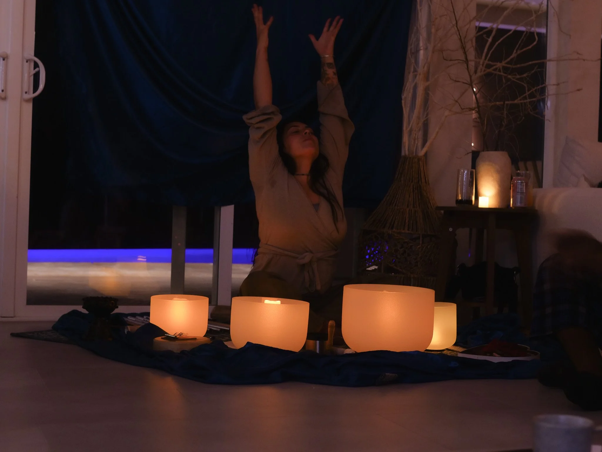 A woman practicing yoga or meditation indoors at night, with her arms raised above her head, surrounded by glowing candles and dim lighting.