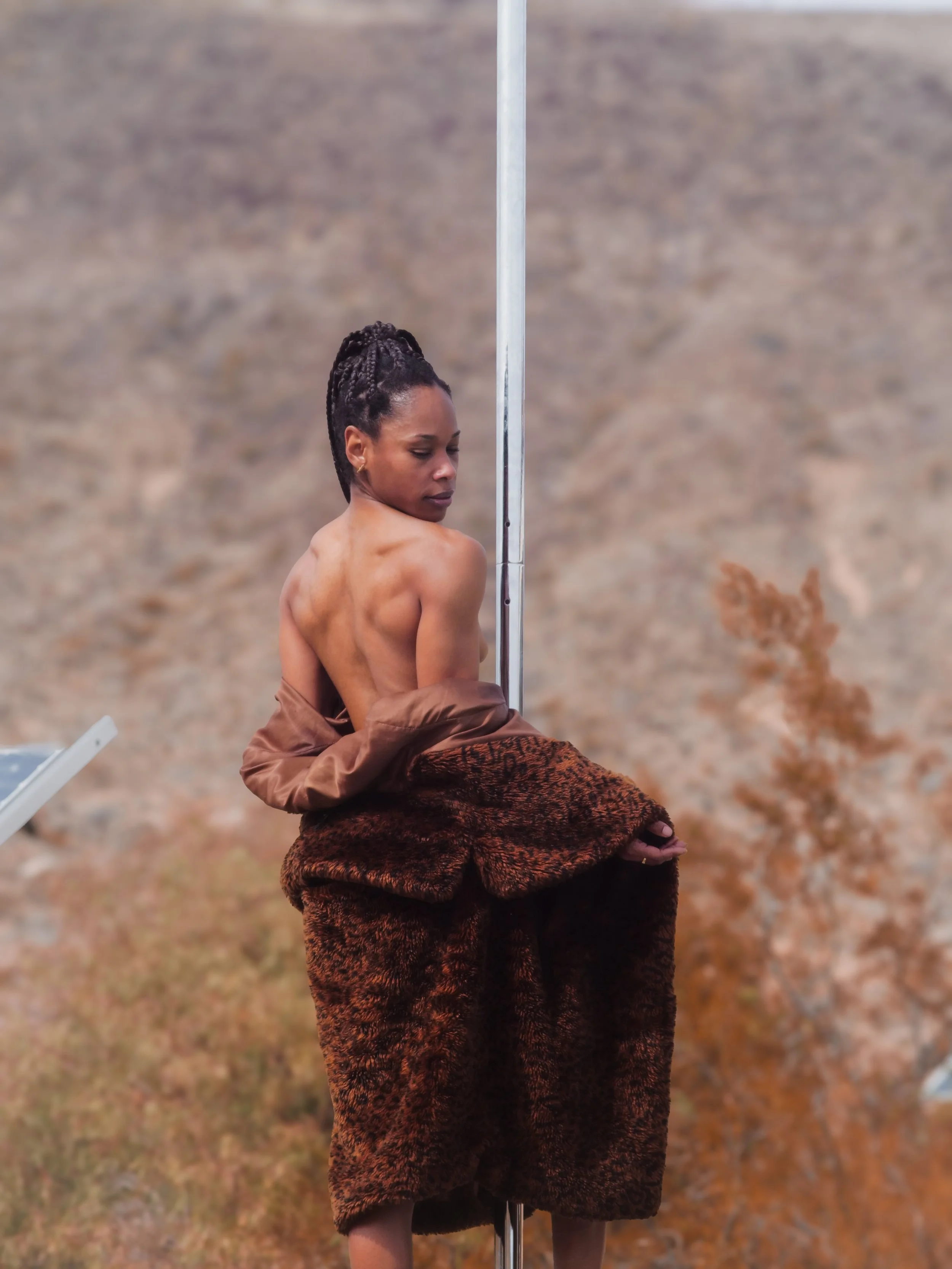 A woman with braided hair sitting outdoors near a metal pole, wearing a brown fur coat around her waist, and a brown jacket. She appears to be topless, facing slightly away with her eyes closed, in a natural landscape background.