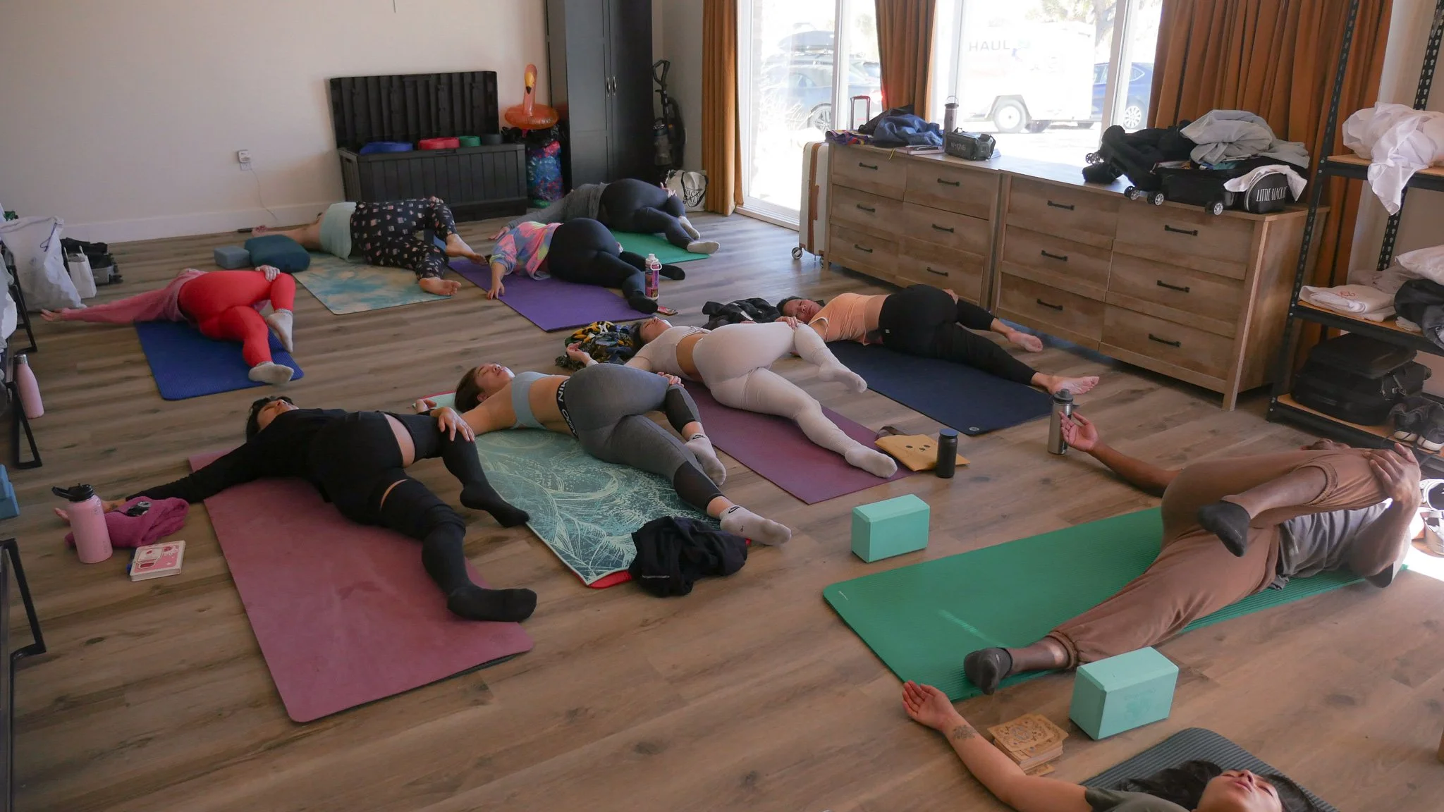 People participating in a prenatal yoga class lying on colorful yoga mats in a studio with wooden floors and large windows.