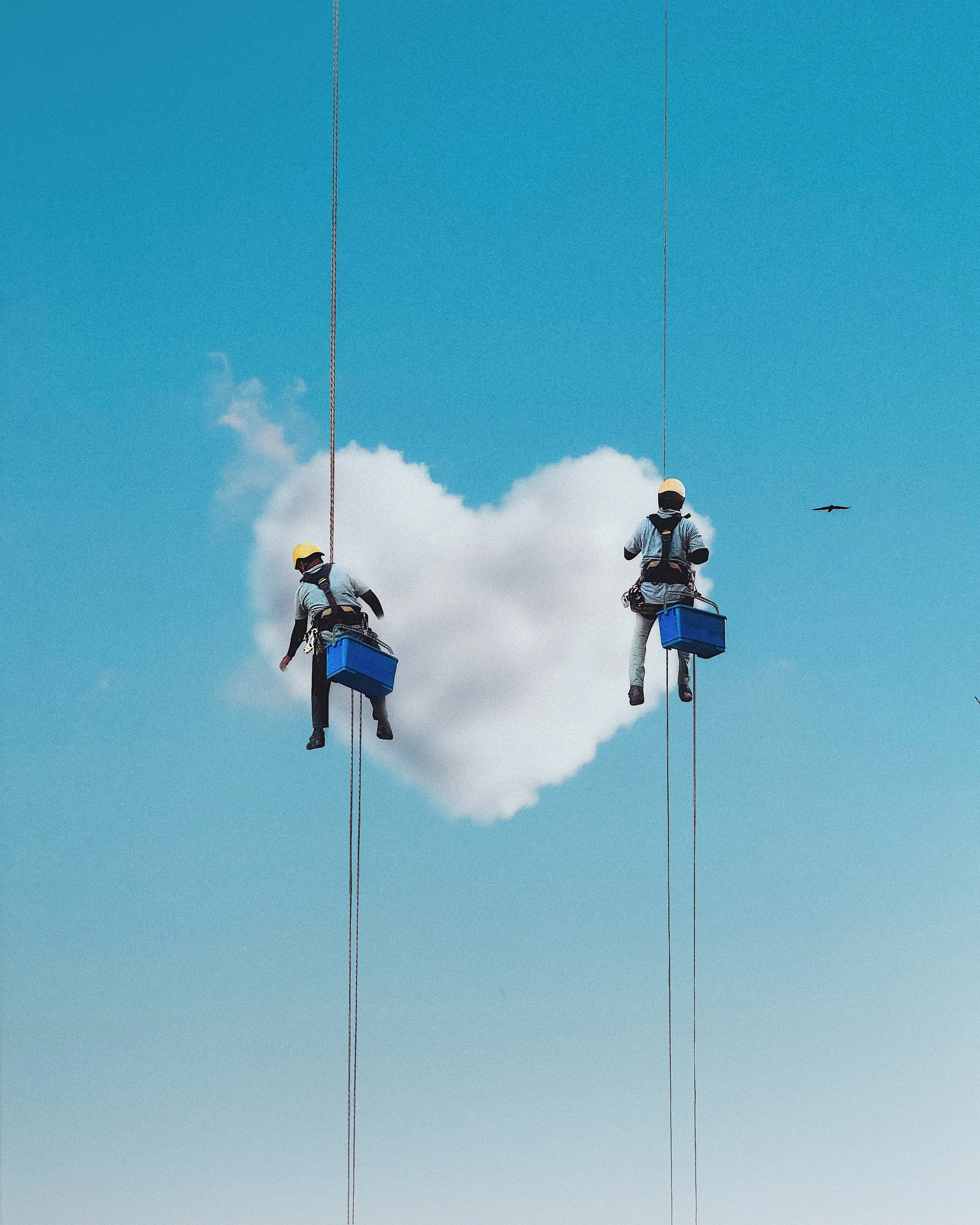 Two workers wearing helmets and safety harnesses are suspended on ropes in front of a large cloud shaped like a heart in the sky, with a bird flying nearby.