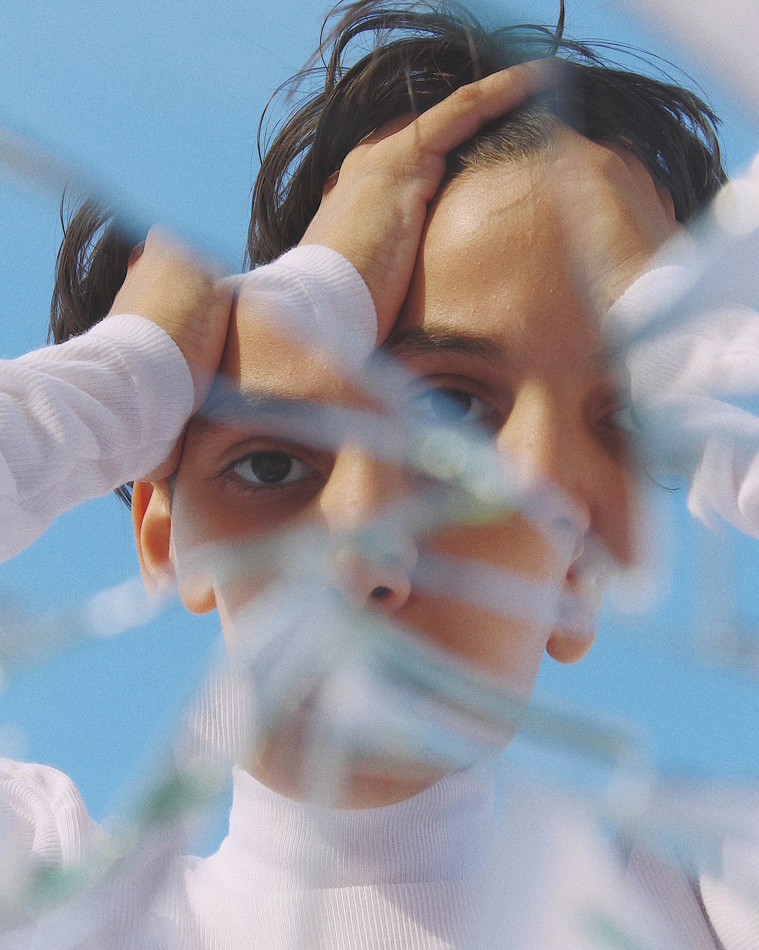 A young person with dark hair partially covering their forehead, looking through blurred white flowers with a clear blue sky in the background.