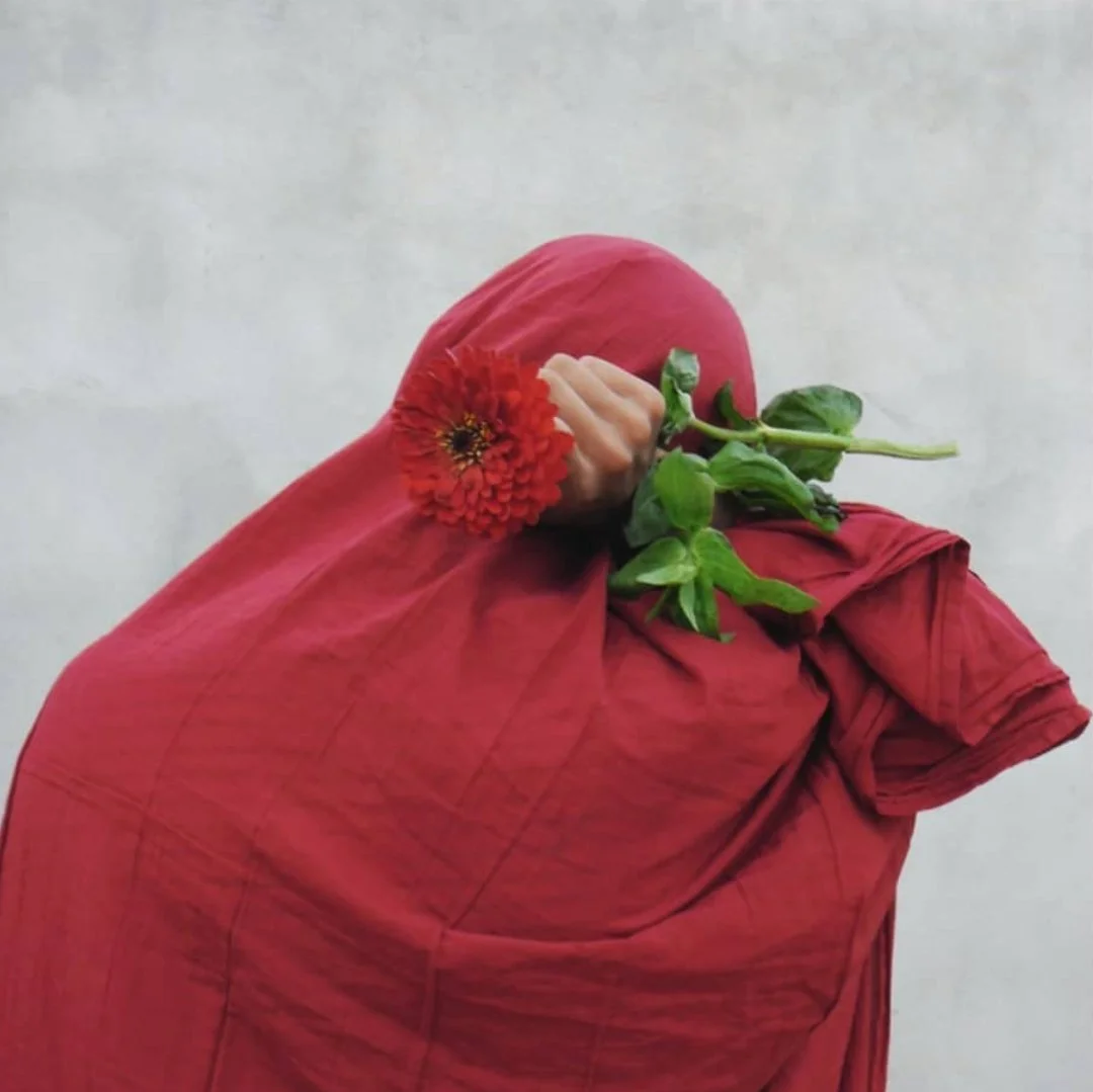 Person wrapped in red cloth holding a red flower and green leaves.