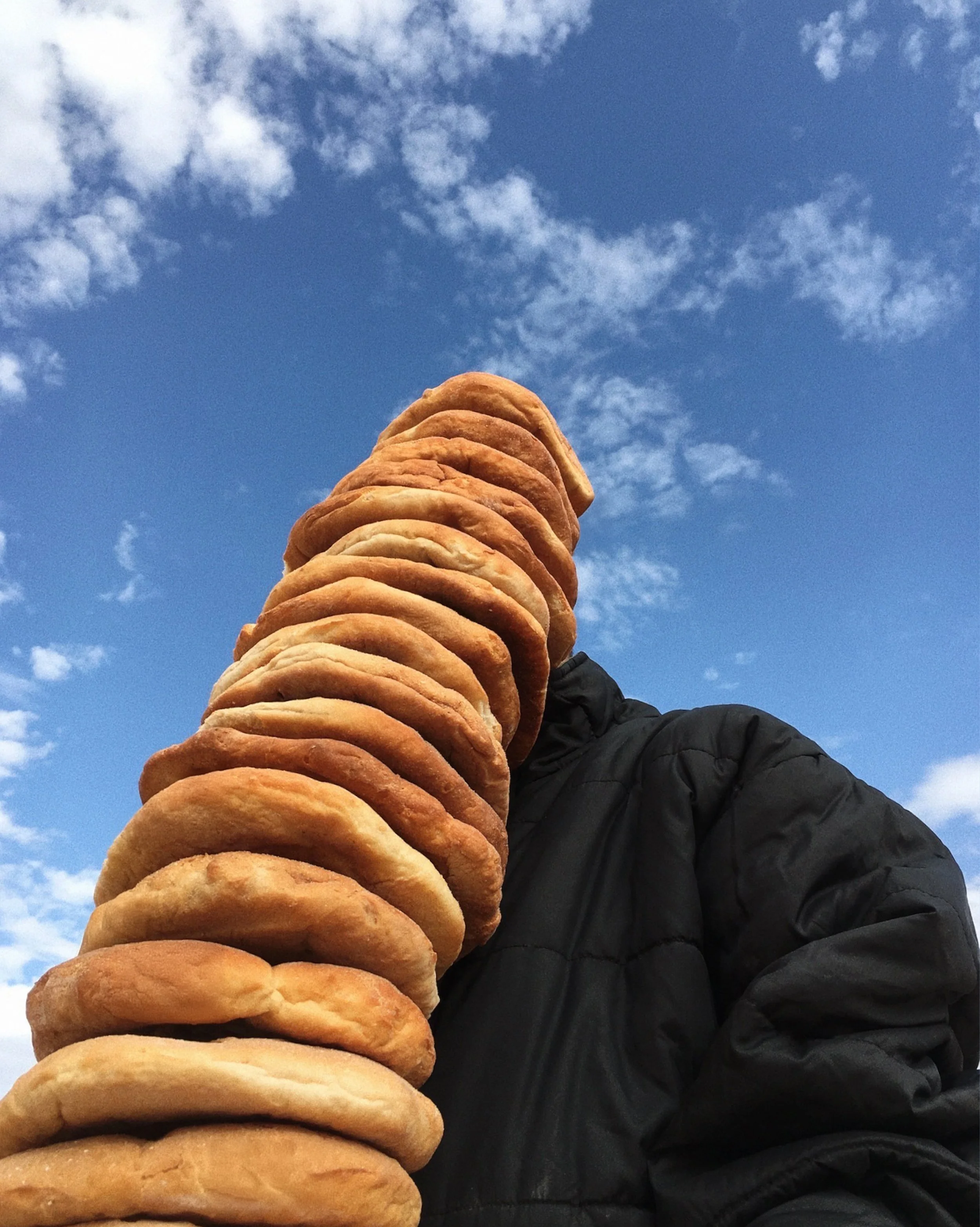 Person holding a tall stack of fried doughnuts outdoors against a blue sky with clouds.