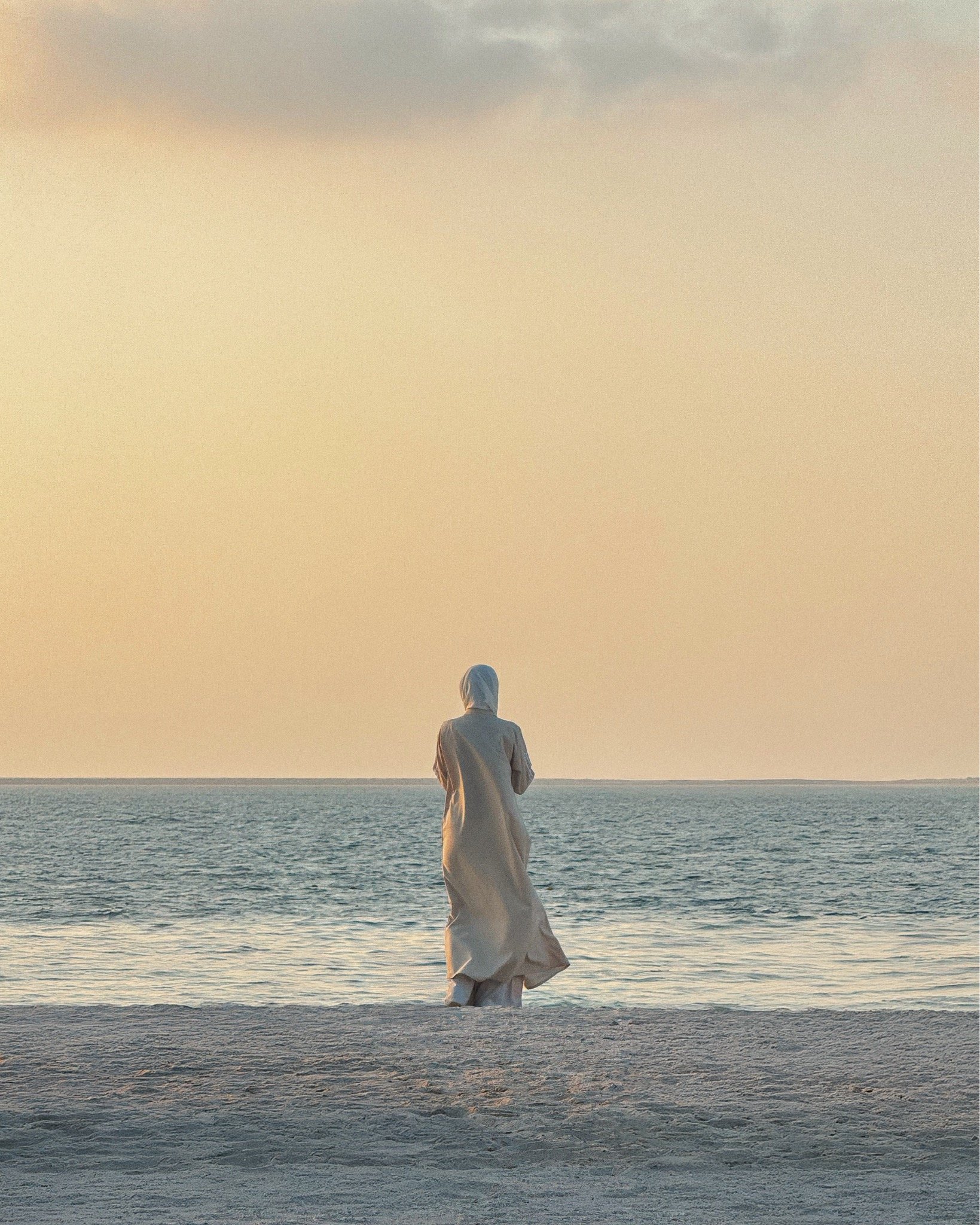A person wearing a long, light-colored cloak and a head covering standing on a sandy beach, facing the ocean during sunset or sunrise.