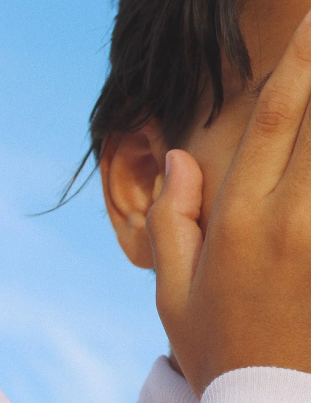 Close-up of a person's hand touching their ear, with dark hair and a light blue background.