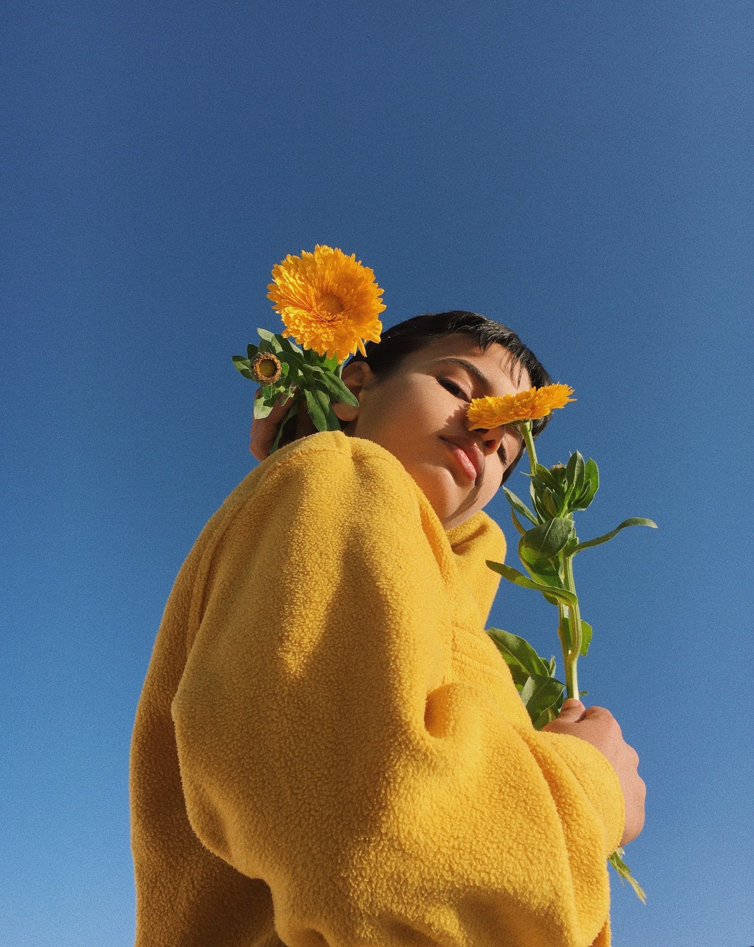 A young woman in a yellow fleece jacket holds yellow flowers and smiles against a clear blue sky.