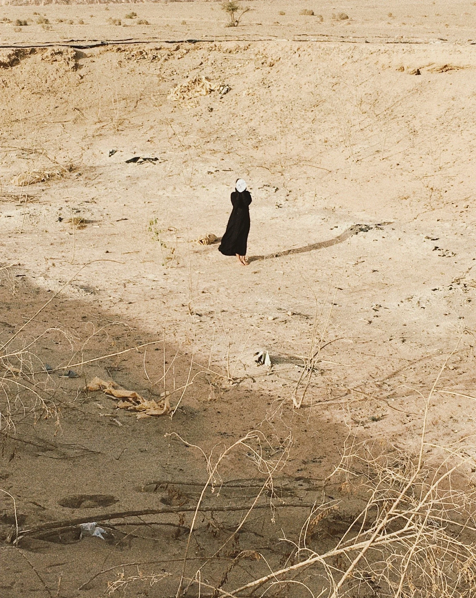 A person in a black dress and white head covering walking alone in a barren, sandy desert landscape with dry bushes and scattered debris.