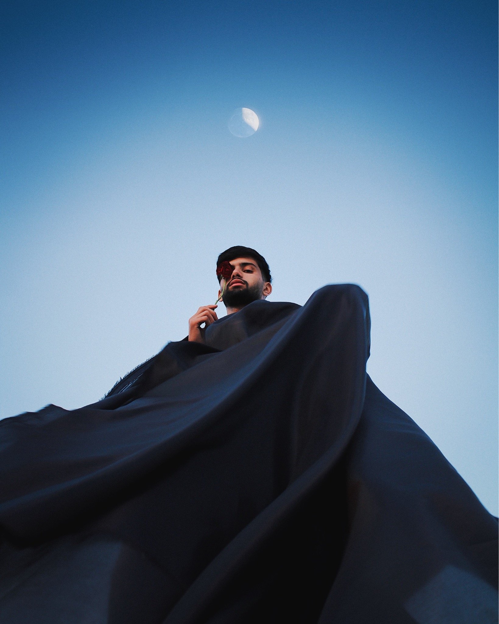 A man in black clothing holding a red flower near his face, looking down at the camera with the moon in the sky above.