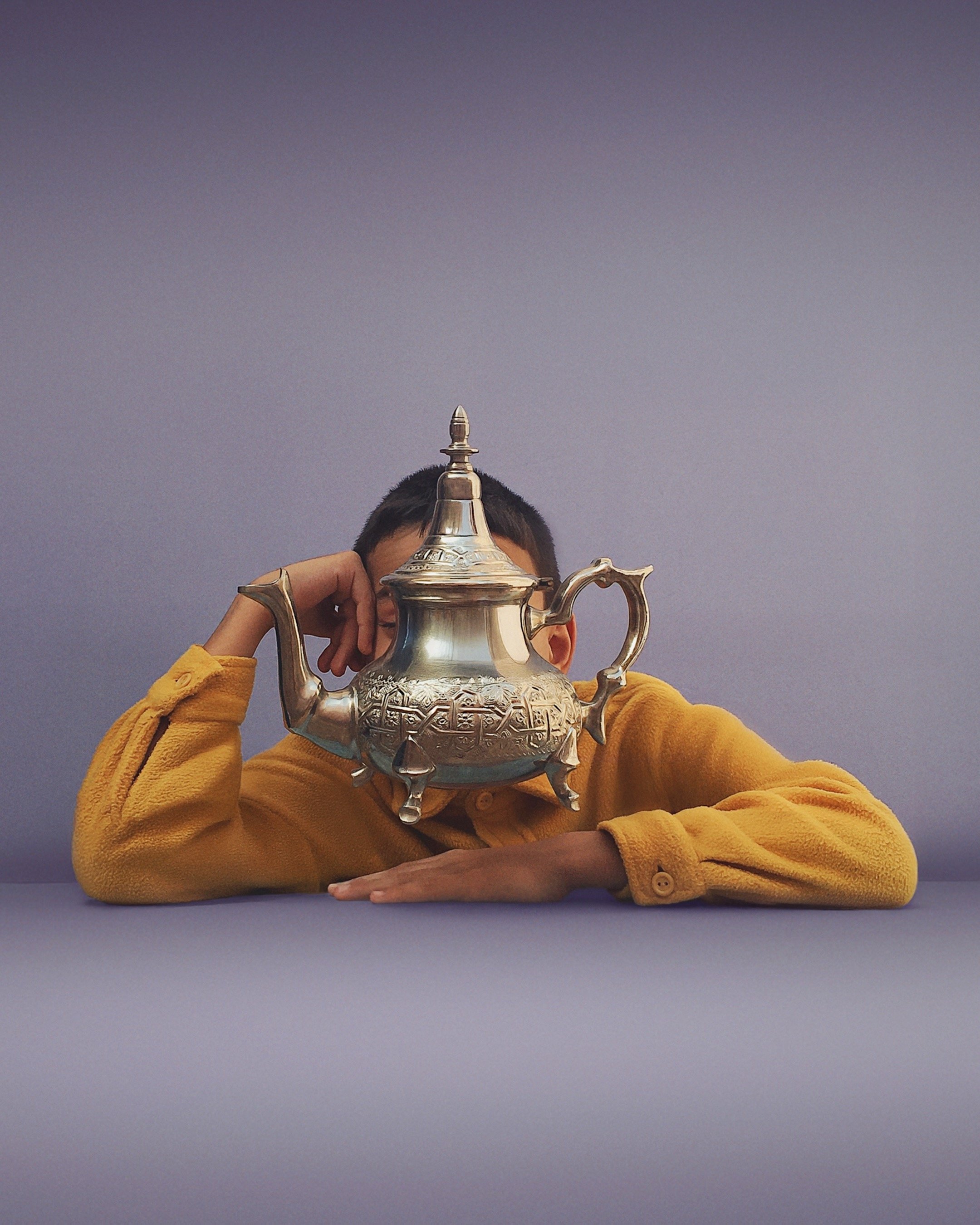 Child in yellow sweater resting on a table with a teapot in front of their face, obscuring it.