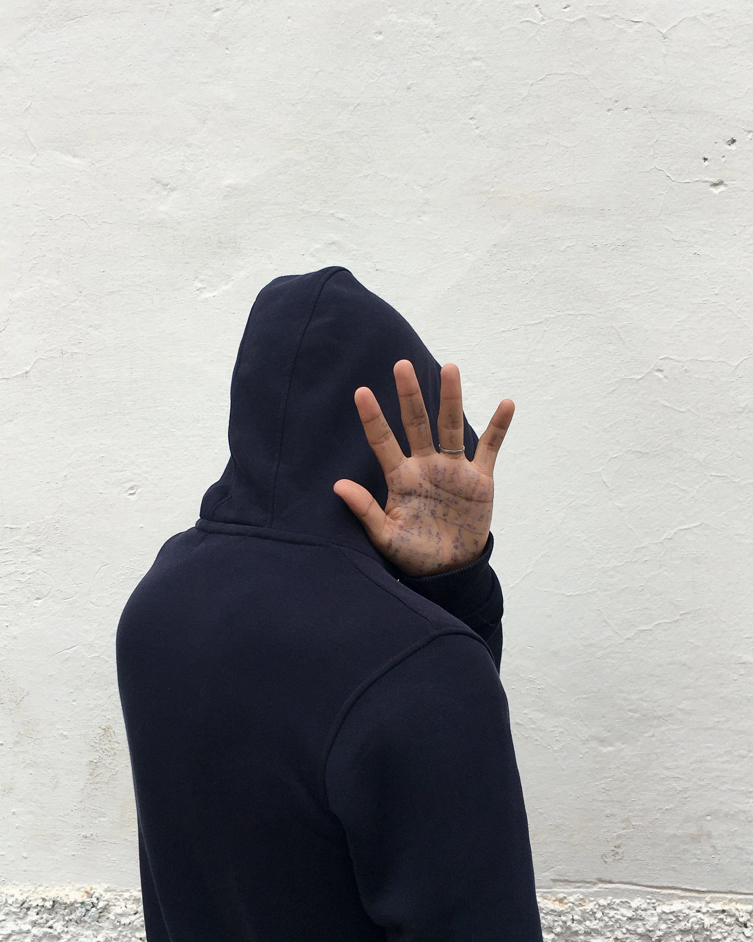 Person wearing a black hoodie with the hood up, covering their face, holding up their hand with intricate purple tattoos on their fingers, standing against a plain, off-white textured wall.