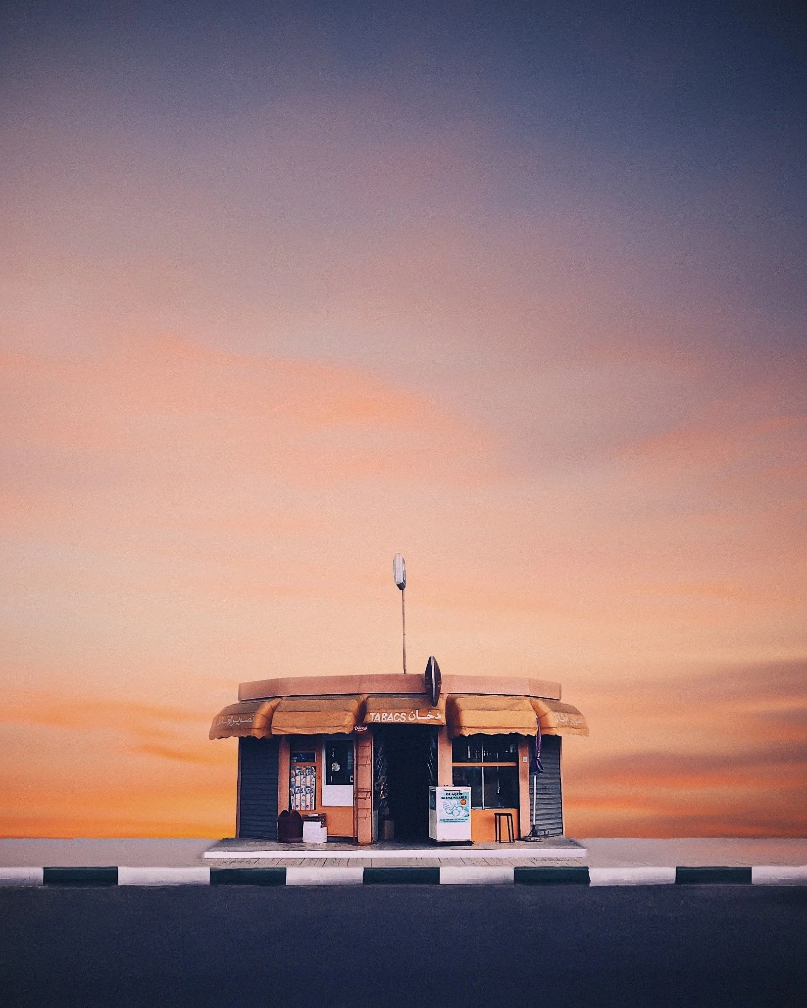 Small kiosk shop with orange awnings and a sign that says 'Tabacs,' located on a sidewalk with a sunset sky background.