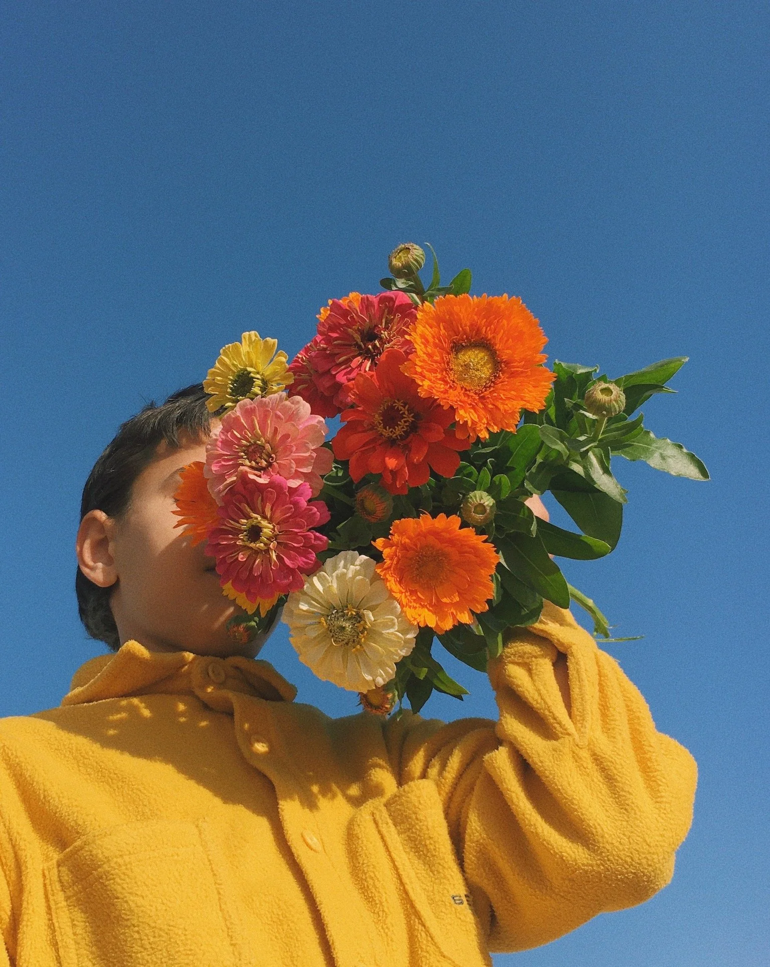 Person in a yellow hoodie holding a bouquet of colorful flowers against a clear blue sky.