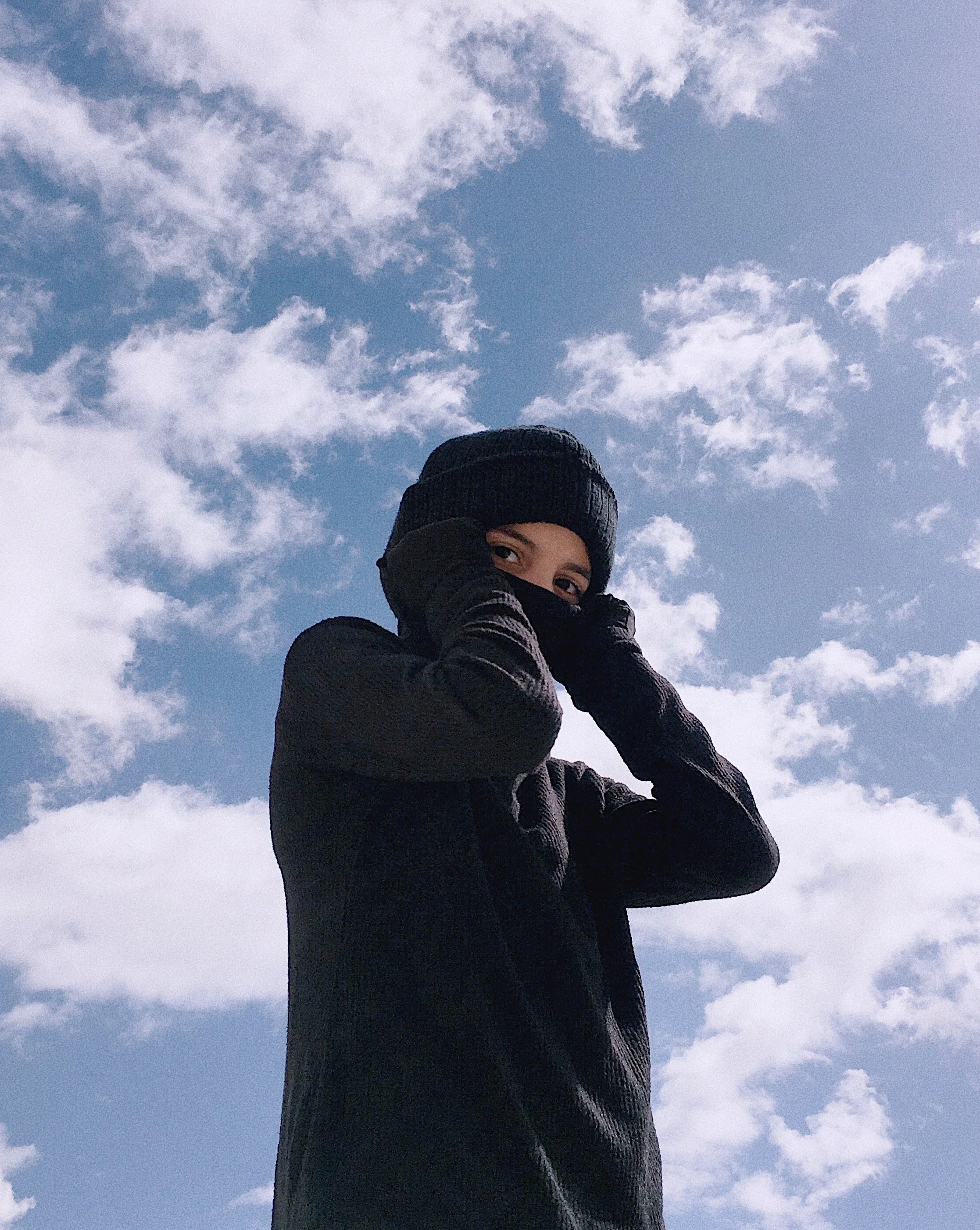 Person dressed in black winter clothing, including a beanie and gloves, standing against a partly cloudy sky.