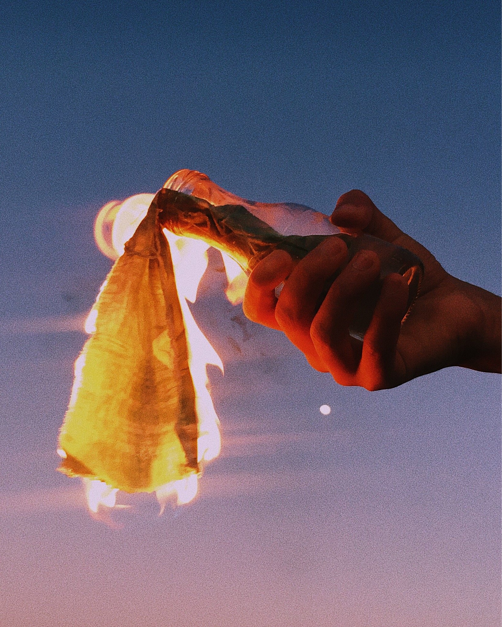 A hand holding a burning piece of paper against a clear evening sky with a visible moon.