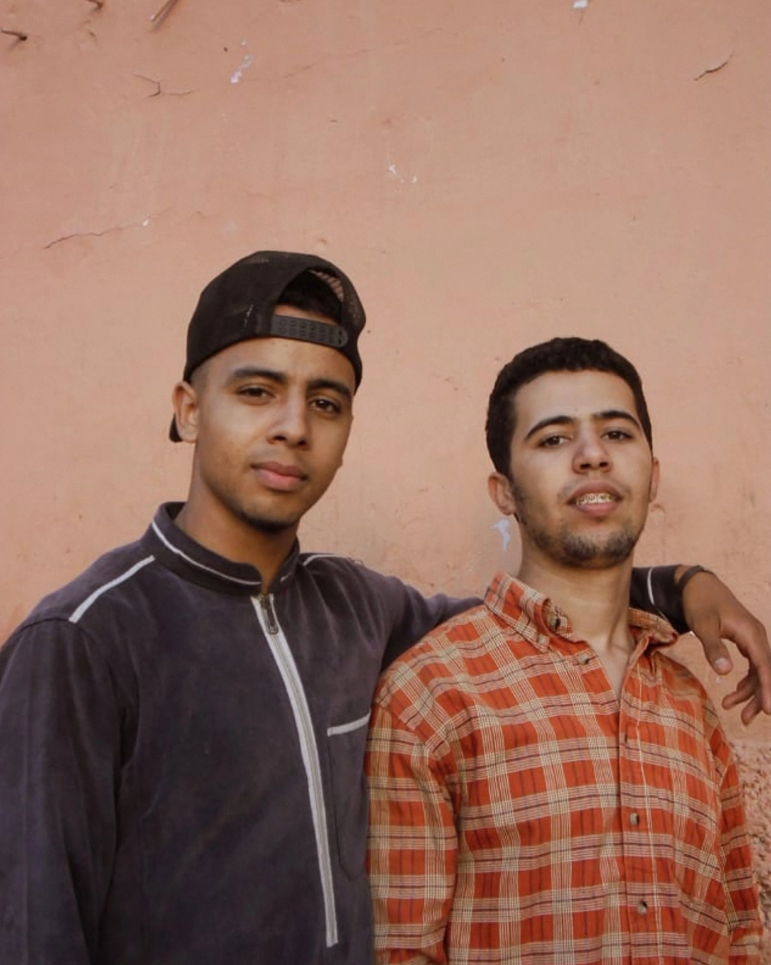 Two young men standing close together in front of a pale pink wall, with one man’s arm around the other's shoulder. One man is wearing a black jacket and a backward baseball cap, while the other is wearing an orange plaid shirt.