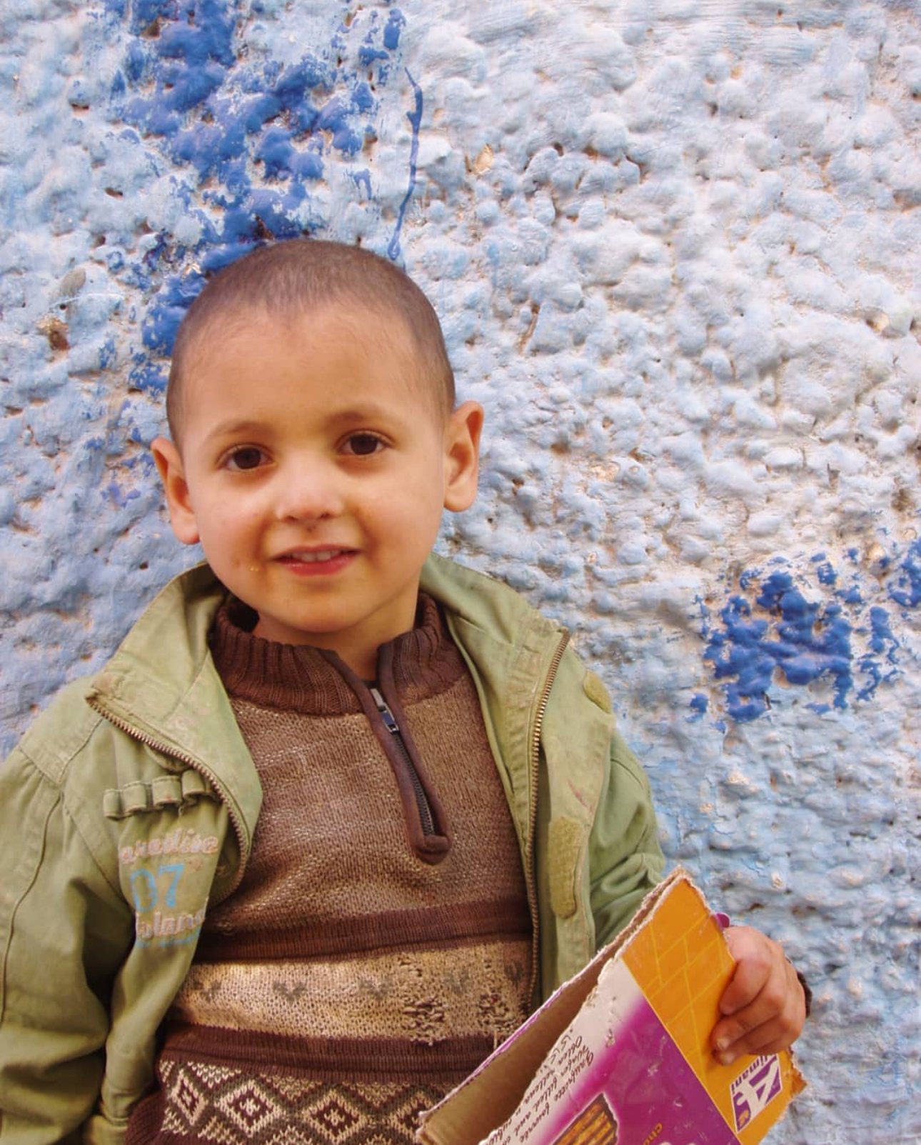 Young boy with a short haircut smiling, wearing a brown sweater and green jacket, holding a torn orange cardboard box, standing in front of a textured white wall with blue paint splatters.