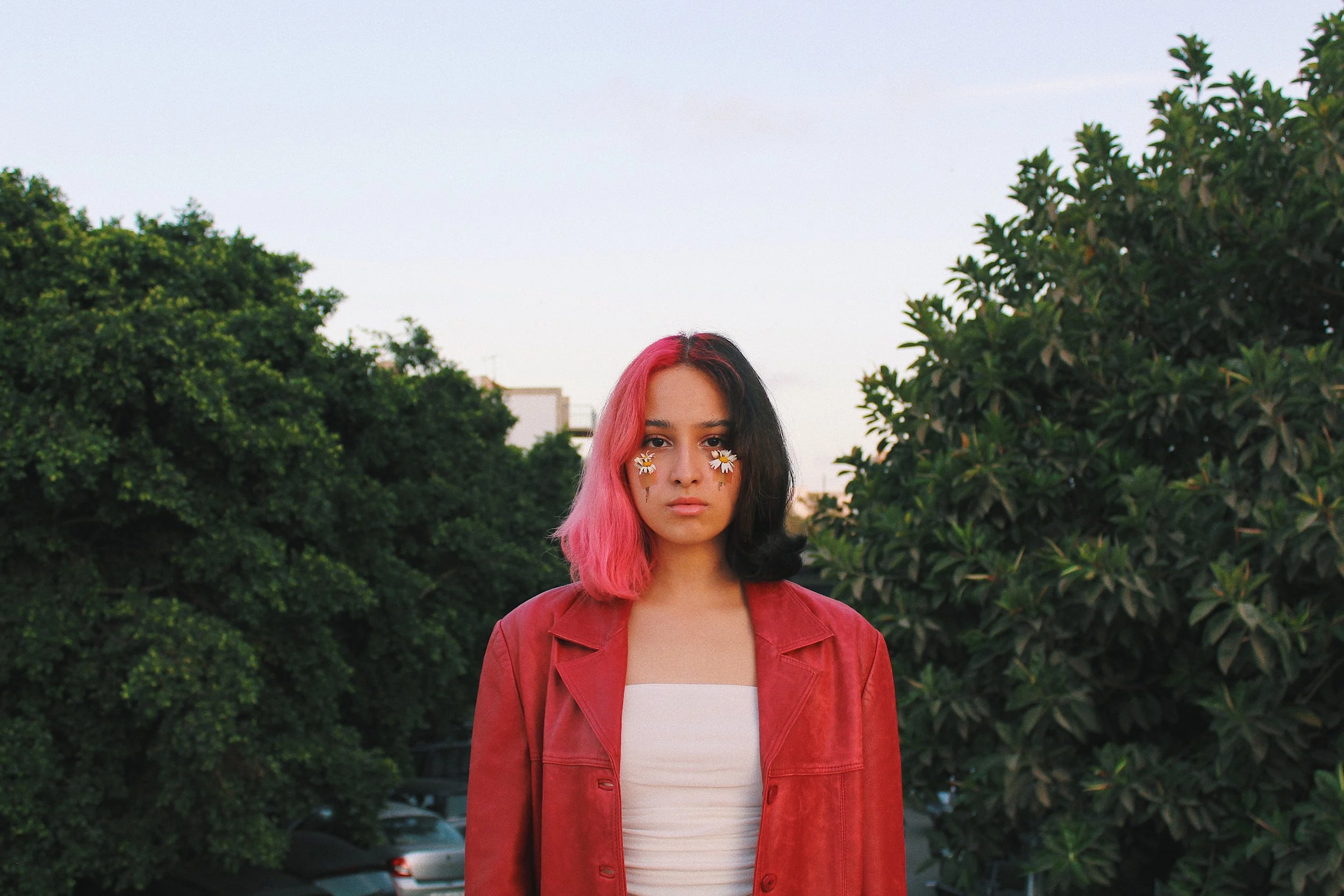 A young woman with half pink and half black hair stands outdoors between green trees, wearing a red leather jacket and a white top, with flower and daisy face decals around her eyes.