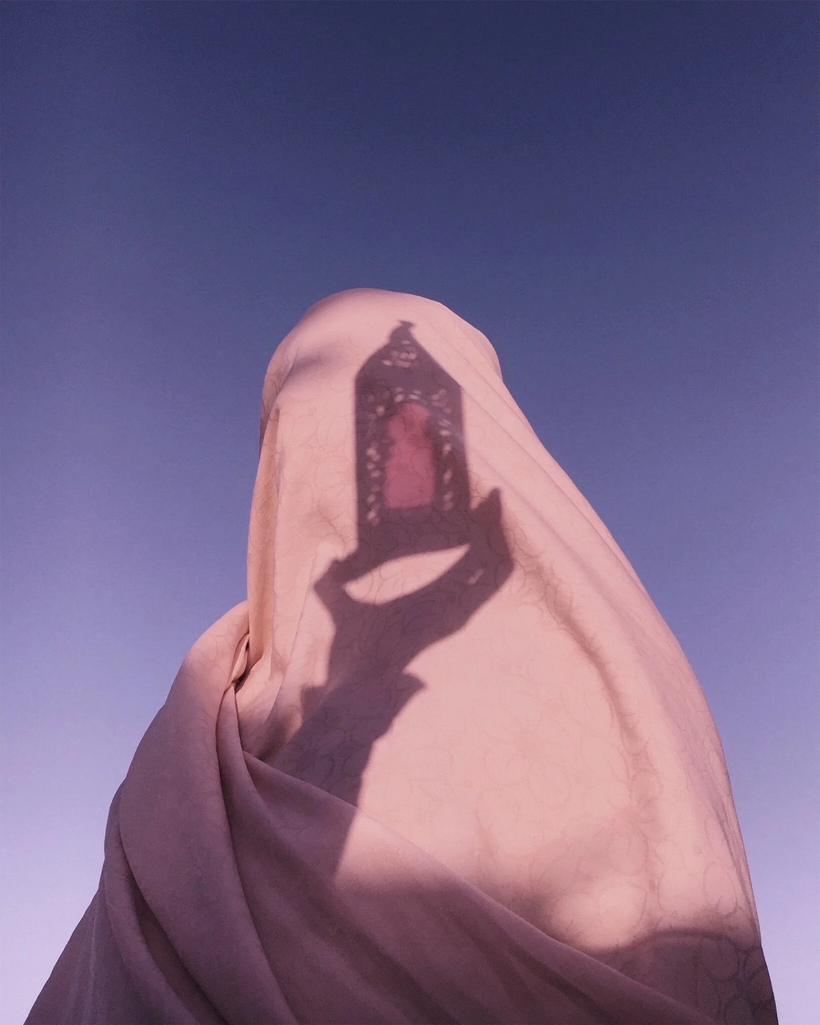 The shadow of a hand holding an ornate lantern is cast onto a pink fabric covering a person's head, with a clear blue sky in the background.