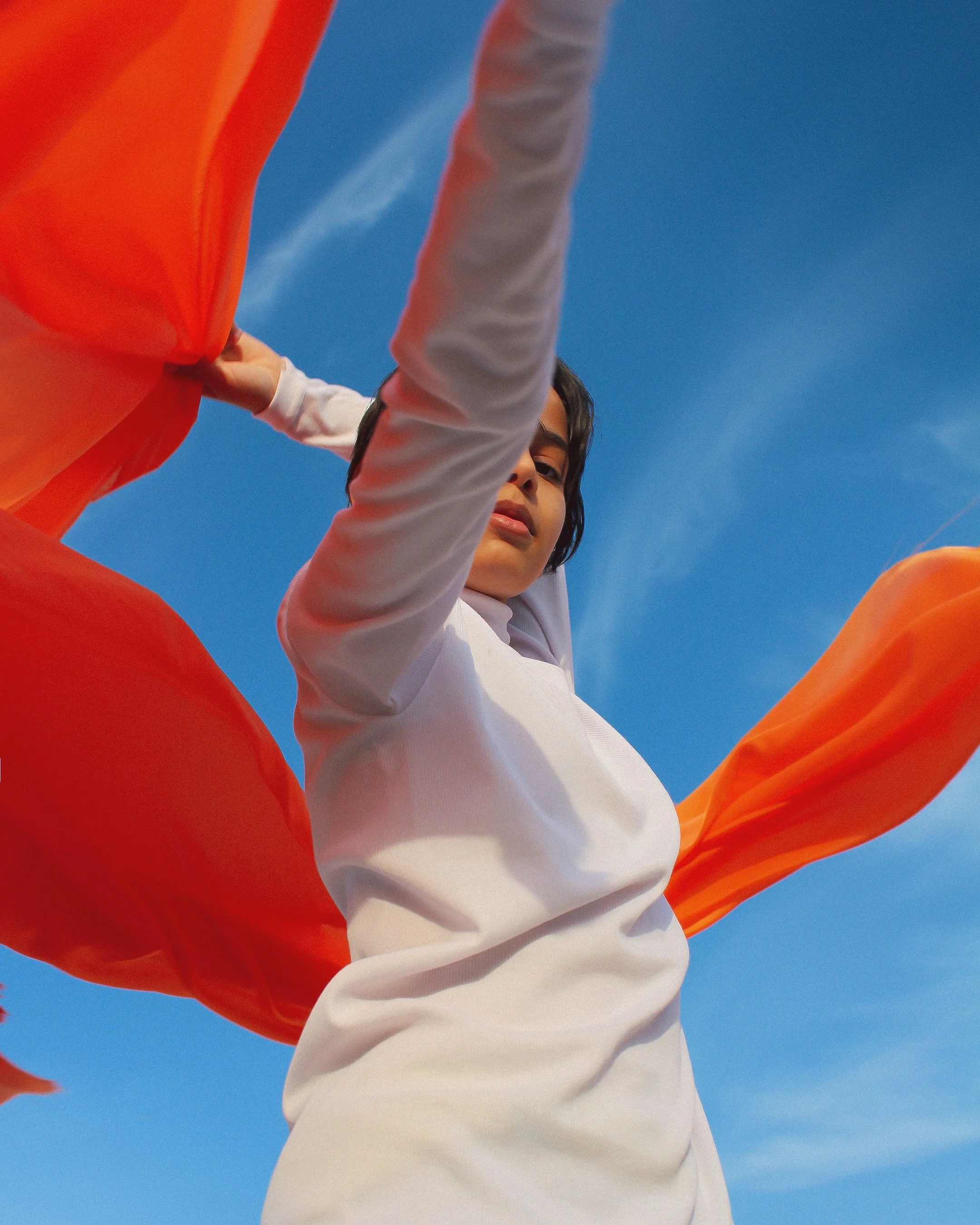 A woman in white reaching upward with a large flowing orange fabric against a clear blue sky.