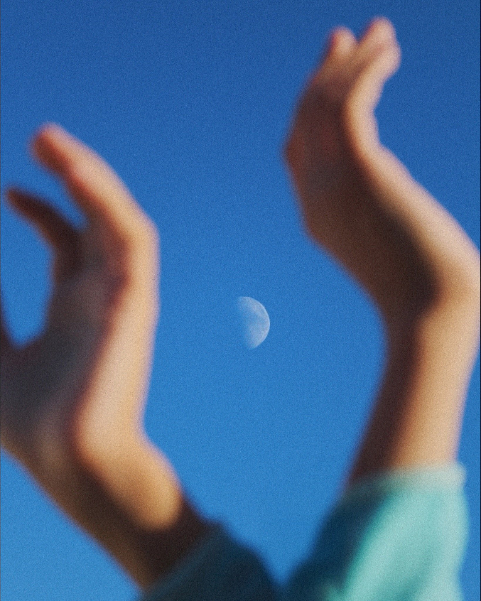 Blurred hands with an open gesture framing the moon in a blue sky.