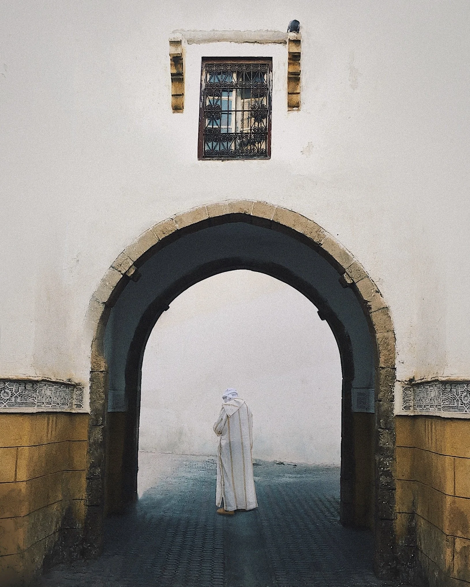 A person dressed in a white traditional robe and head covering stands under an arched gateway with a white wall and a small window above.