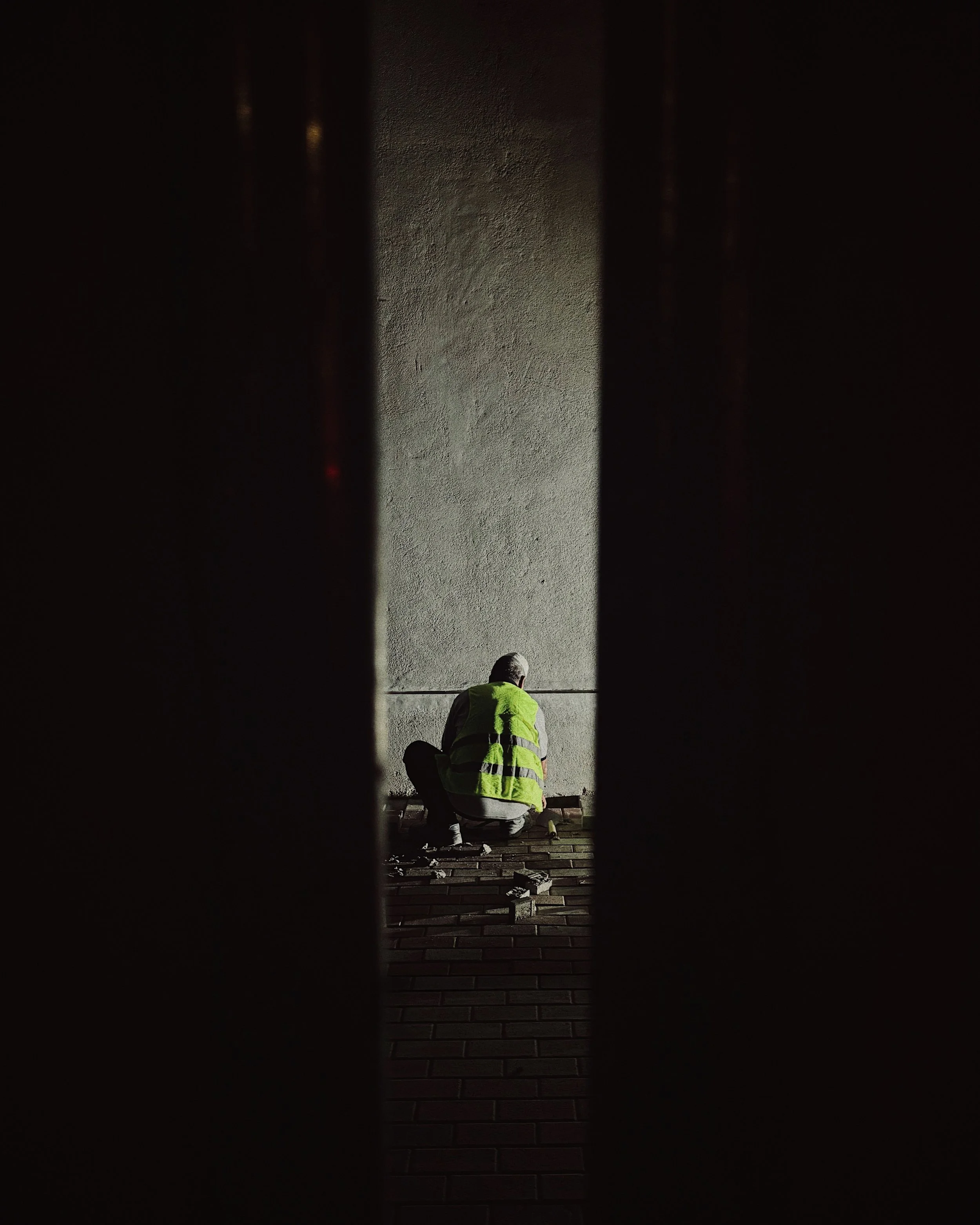Person in a yellow safety vest crouching and working on a wall, viewed through a narrow gap between two dark walls.