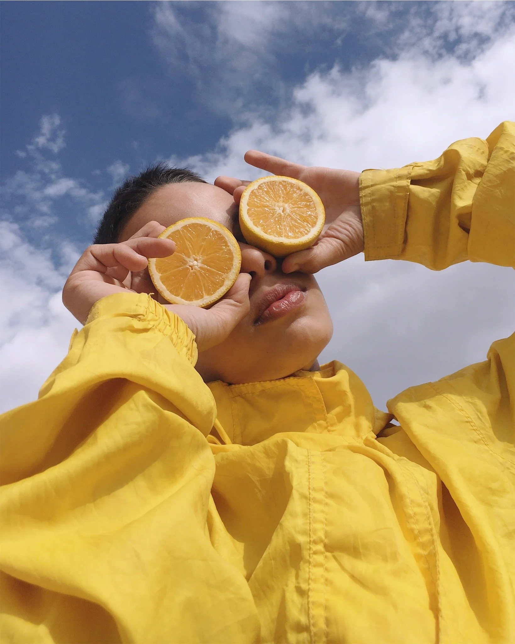 A person in a yellow jacket holding two lemon halves in front of their face against a partly cloudy sky.