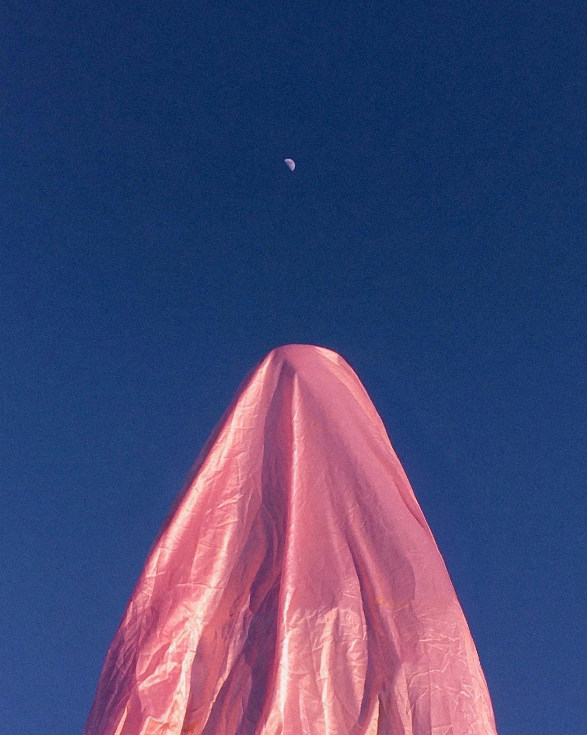 A pink fabric cover or tarp draped over an object, with a clear blue sky in the background and the moon visible at the top center of the image.