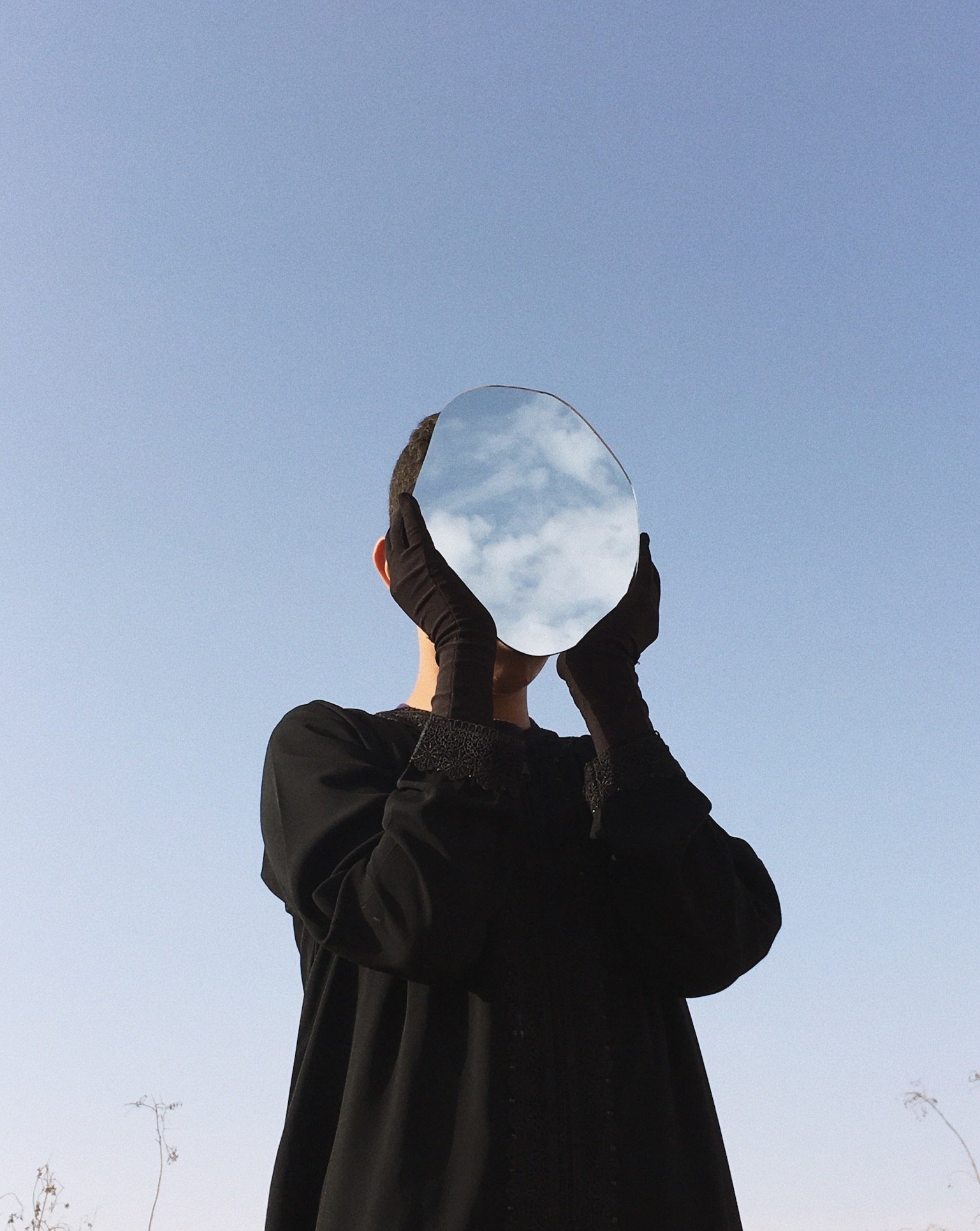 Person wearing black gloves holding a round mirror reflecting the cloudy sky, standing outdoors against a clear blue sky.
