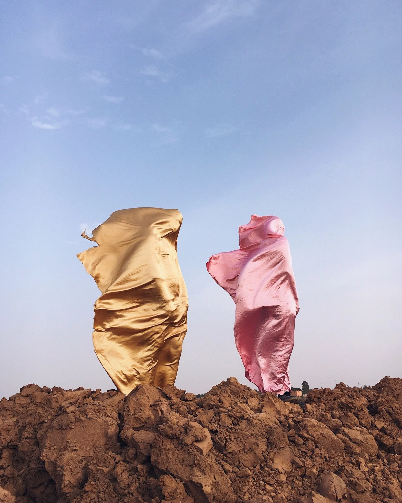 Two large fabrics, one gold and one pink, appear to be floating or waving in the wind above rocky ground against a blue sky.