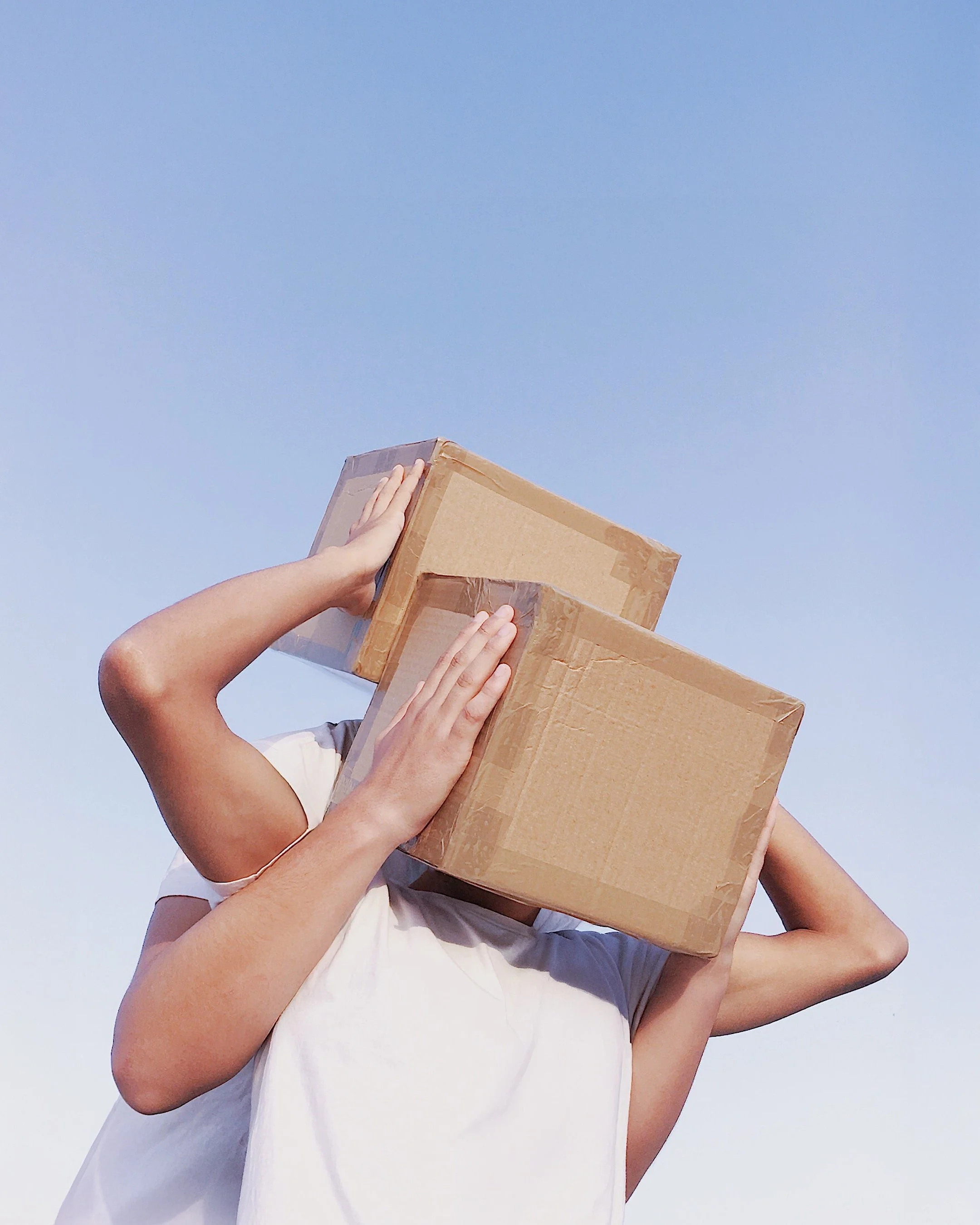 Person carrying two cardboard boxes on their shoulders against a clear blue sky.