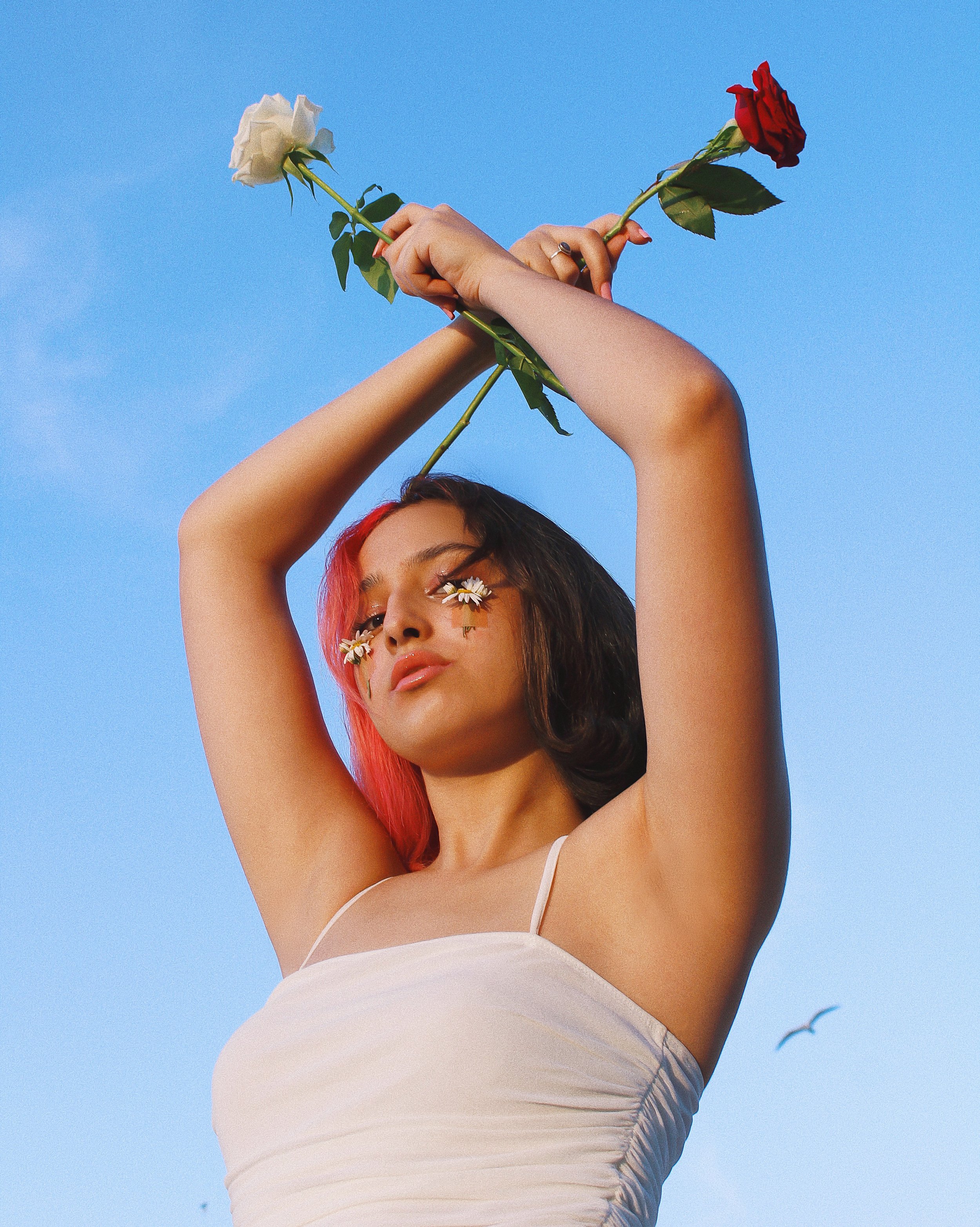 Young woman with flowers on her face, holding two roses in the air against a blue sky.