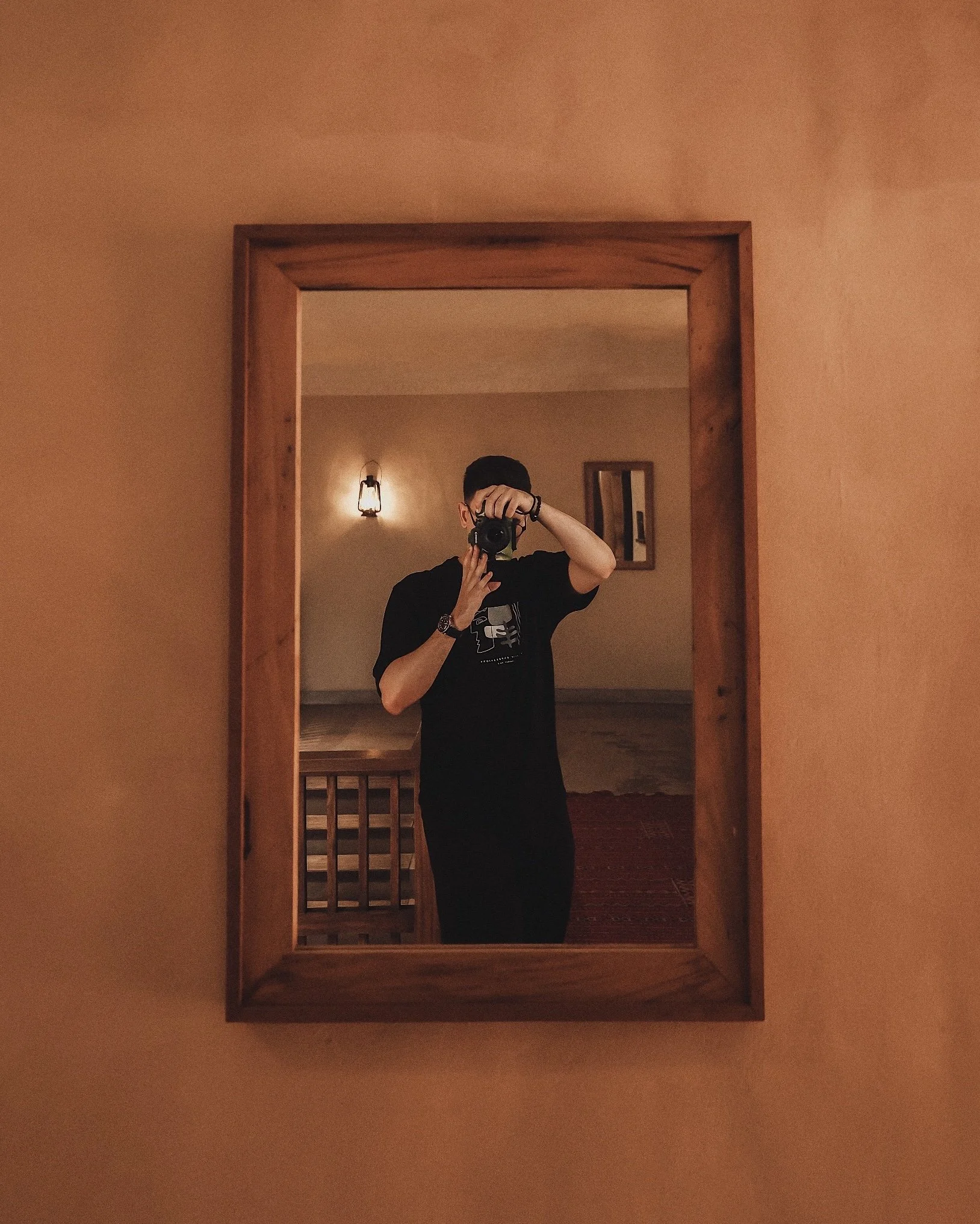 Person taking a photo of themselves in a mirror. The person is standing in a room with beige walls, and a wall sconce light and framed picture are visible in the background.